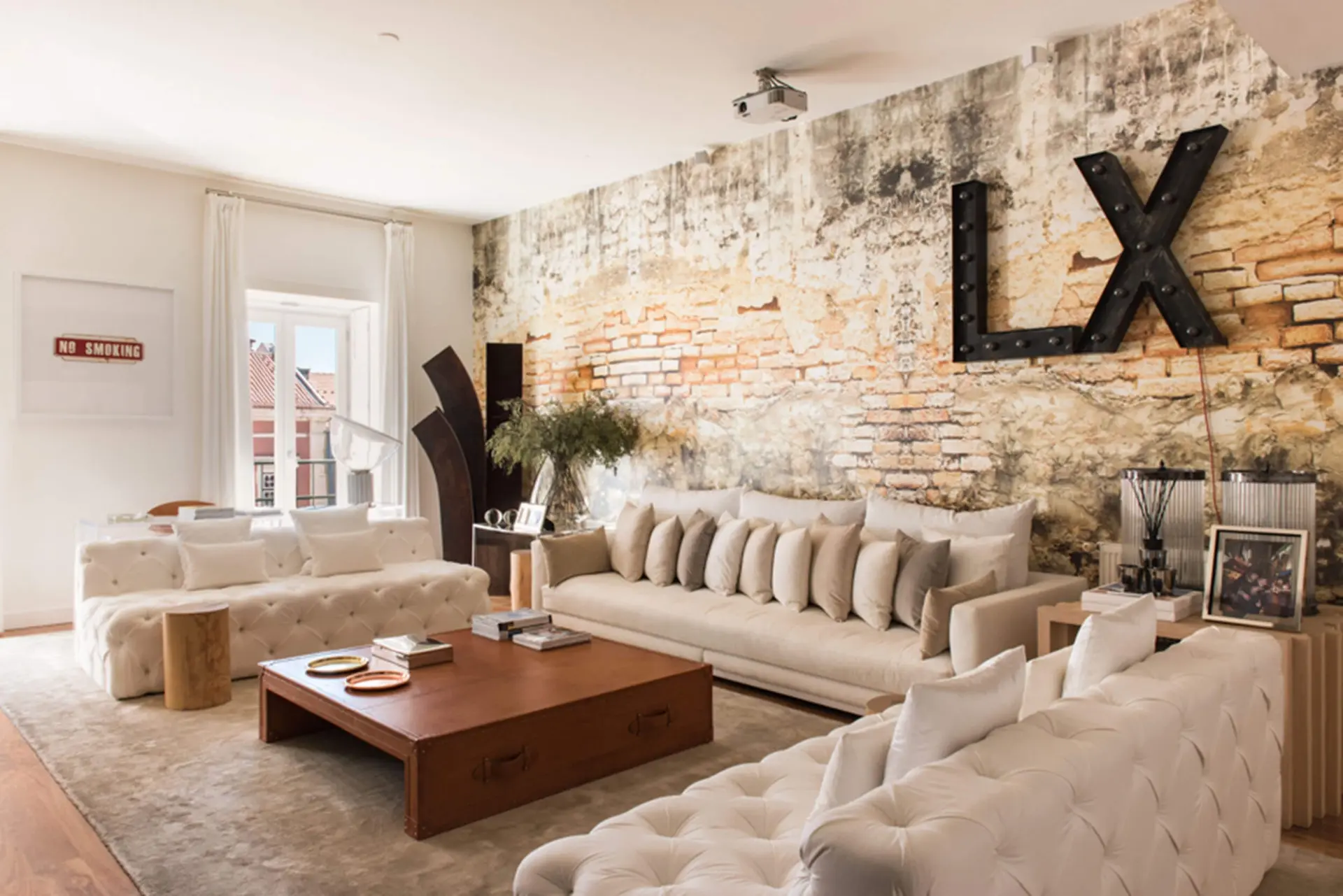 Exposed-brick wall with large metal letters, tufted cream sofas and a campaign-trunk wood coffee table — luxury living room