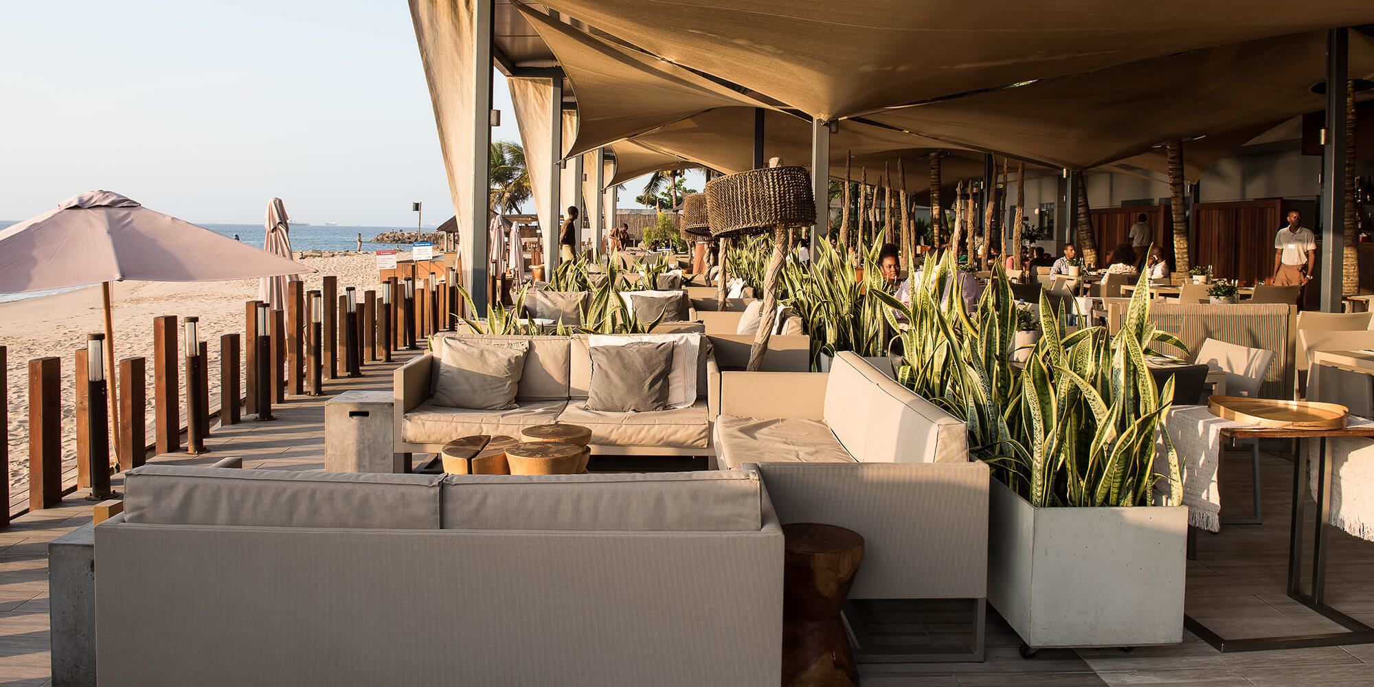 Esplanada de café de praia com sofás em outdoor, plantas tropicais e pérgula de lona com vista para o oceano — projeto de design de interiores em Luanda, Angola, Atelier Renata Santos Machado
