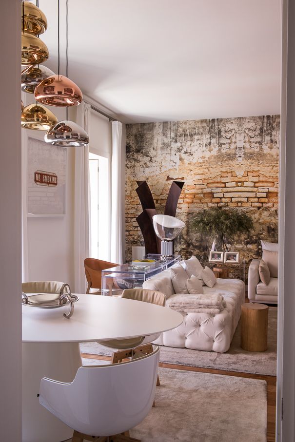 Sala de estar e jantar com sofá claro, mesa branca, candeeiros suspensos metálicos e parede com textura de tijolo aparente.