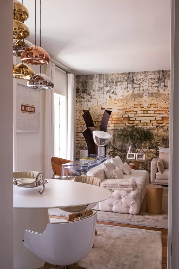 Sala de estar e jantar com sofá claro, mesa branca, candeeiros suspensos metálicos e parede com textura de tijolo aparente.