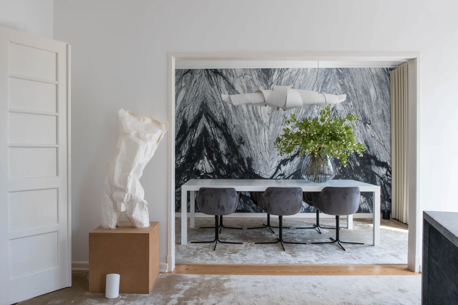 Sala de jantar contemporânea em Lisboa com mesa branca minimalista, cadeiras estofadas cinza, escultura decorativa e parede em mármore preto e branco.