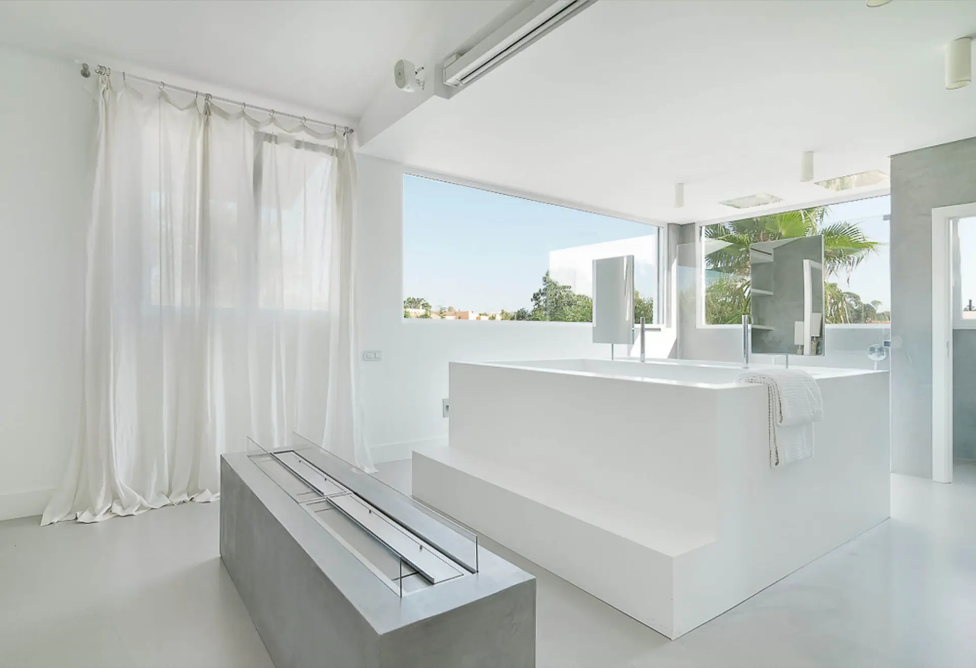 White Corian tub and basin on a concrete base with an ethanol fireplace, linen curtains and a garden view — luxury bathroom