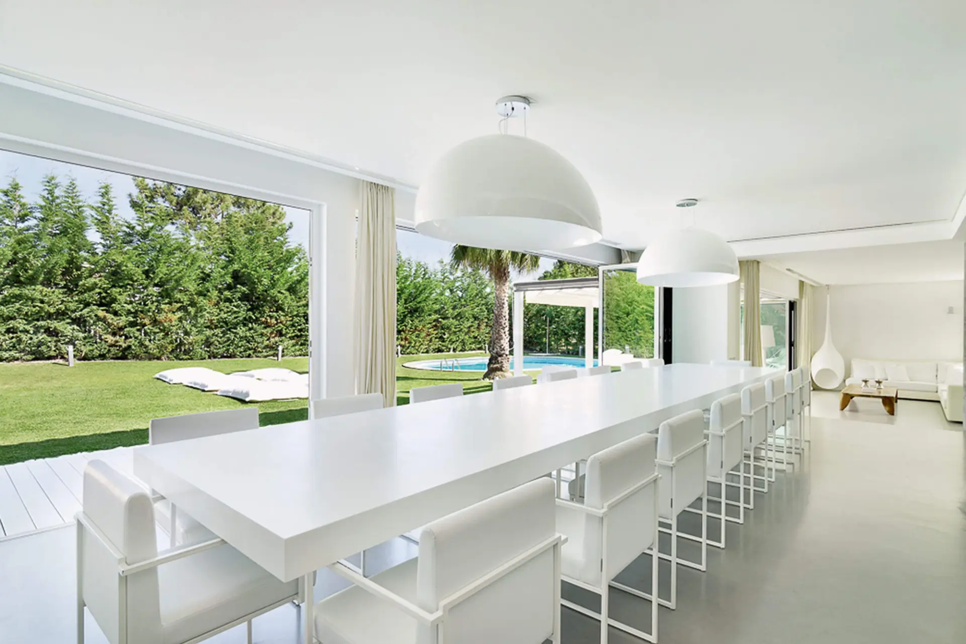 Long white table, white chairs and a pair of oversized white dome pendants by a glass wall to a pool garden — luxury dining room