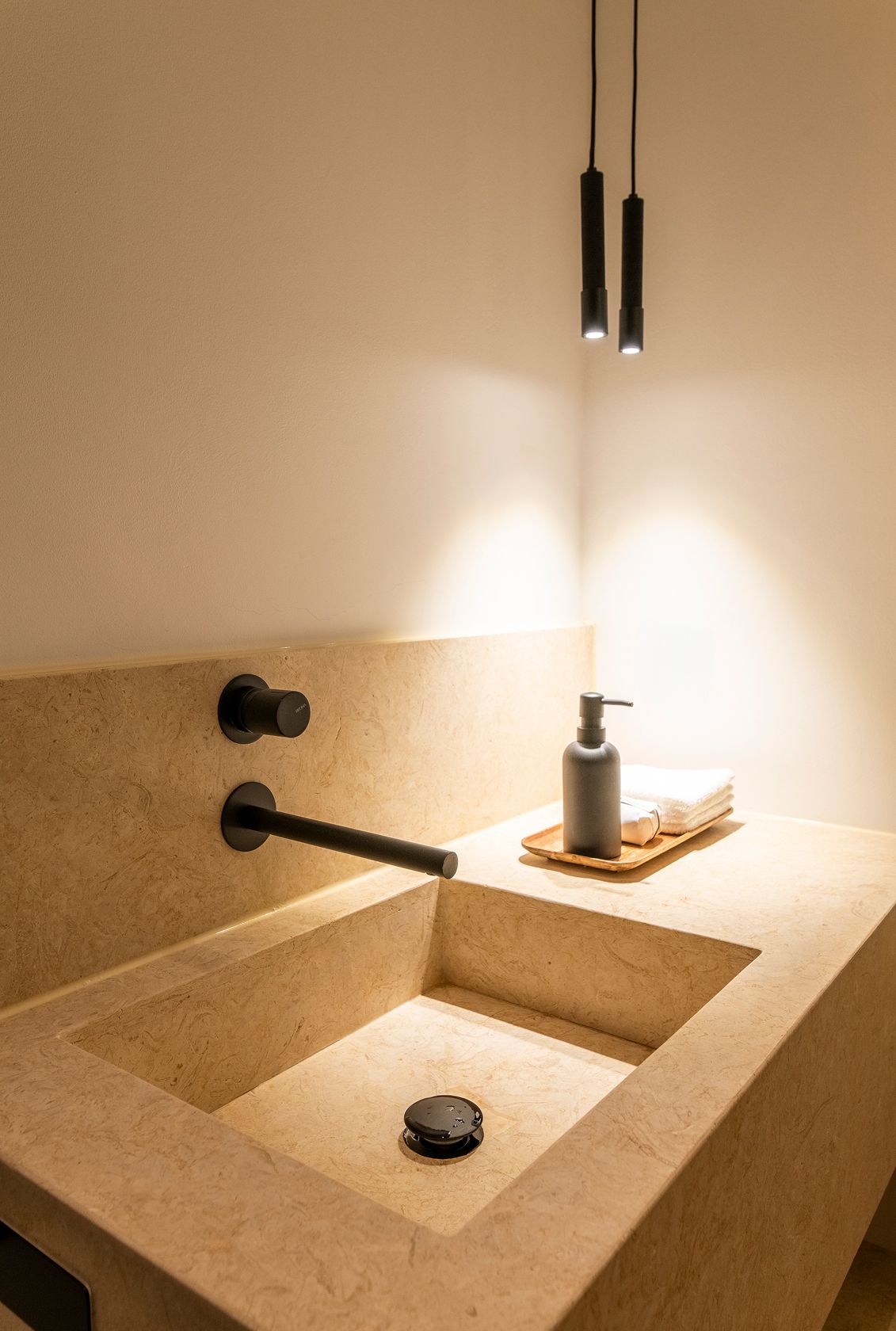 Beige limestone basin, matte black wall tap and slim black tube pendants — luxury décor details