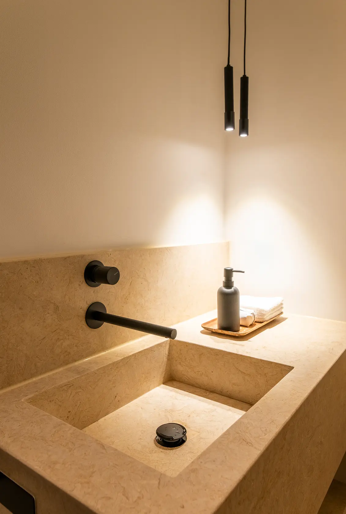 Beige limestone basin, matte black wall tap and slim black tube pendants — luxury décor details
