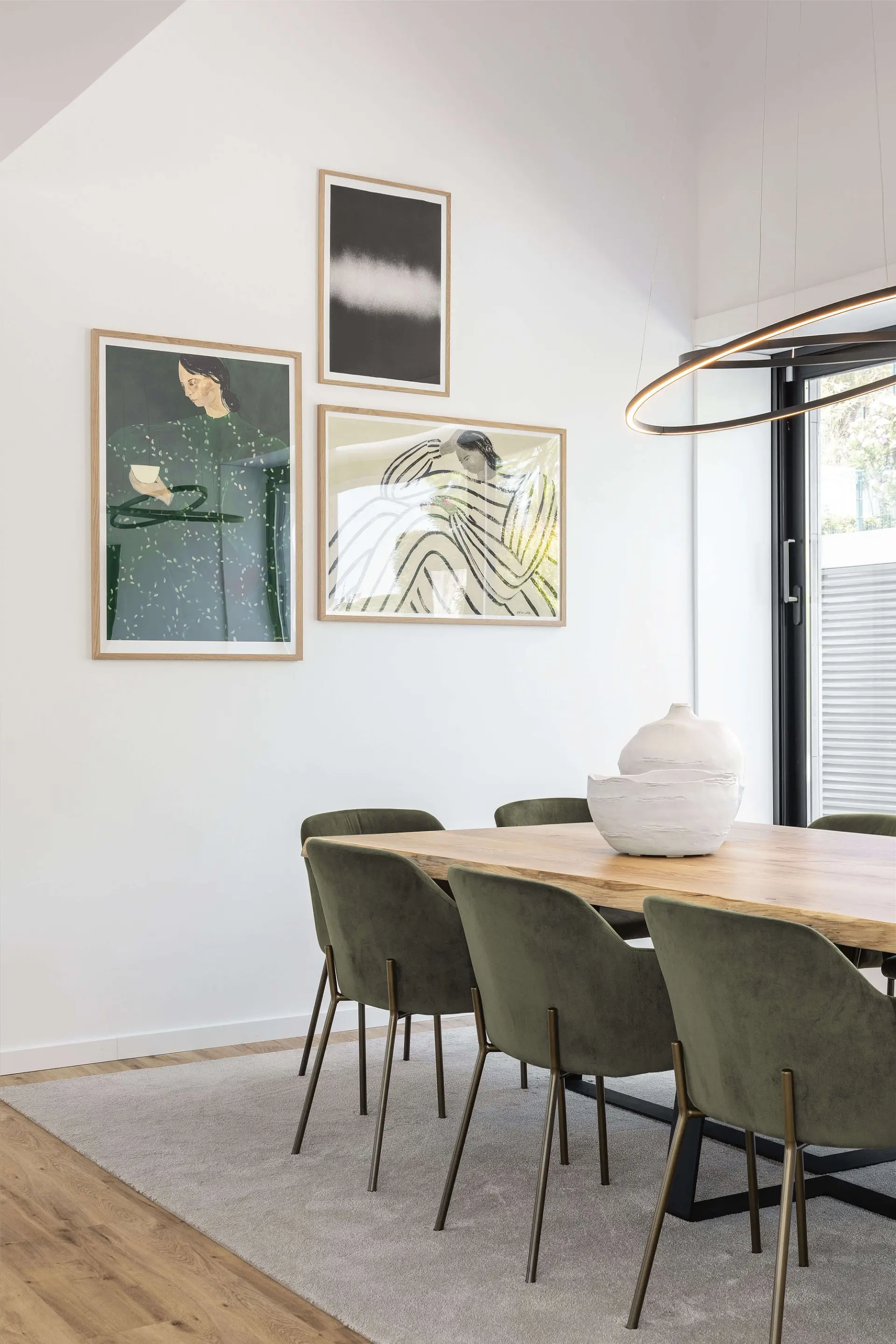 Oak table, olive-green velvet chairs on brass legs and a ring halo pendant below a gallery wall of framed art — luxury dining room