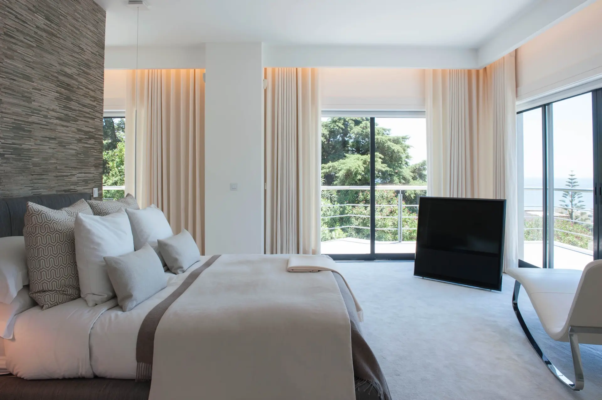 Grey upholstered headboard, stacked stone feature wall and white chaise longue with sea view — luxury master suite design