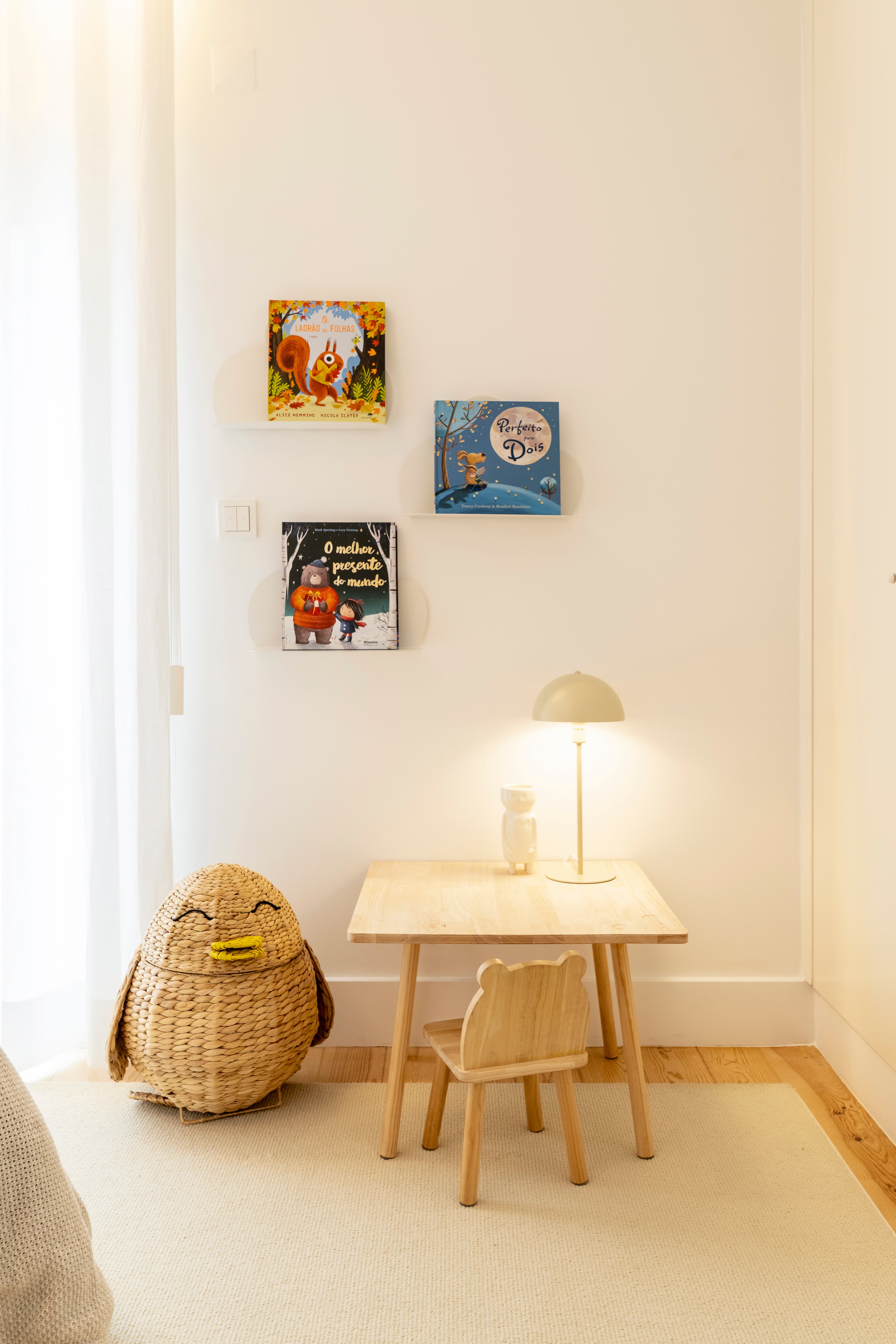 Zona de leitura infantil com mesa e cadeira em madeira, candeeiro e livros expostos na parede.