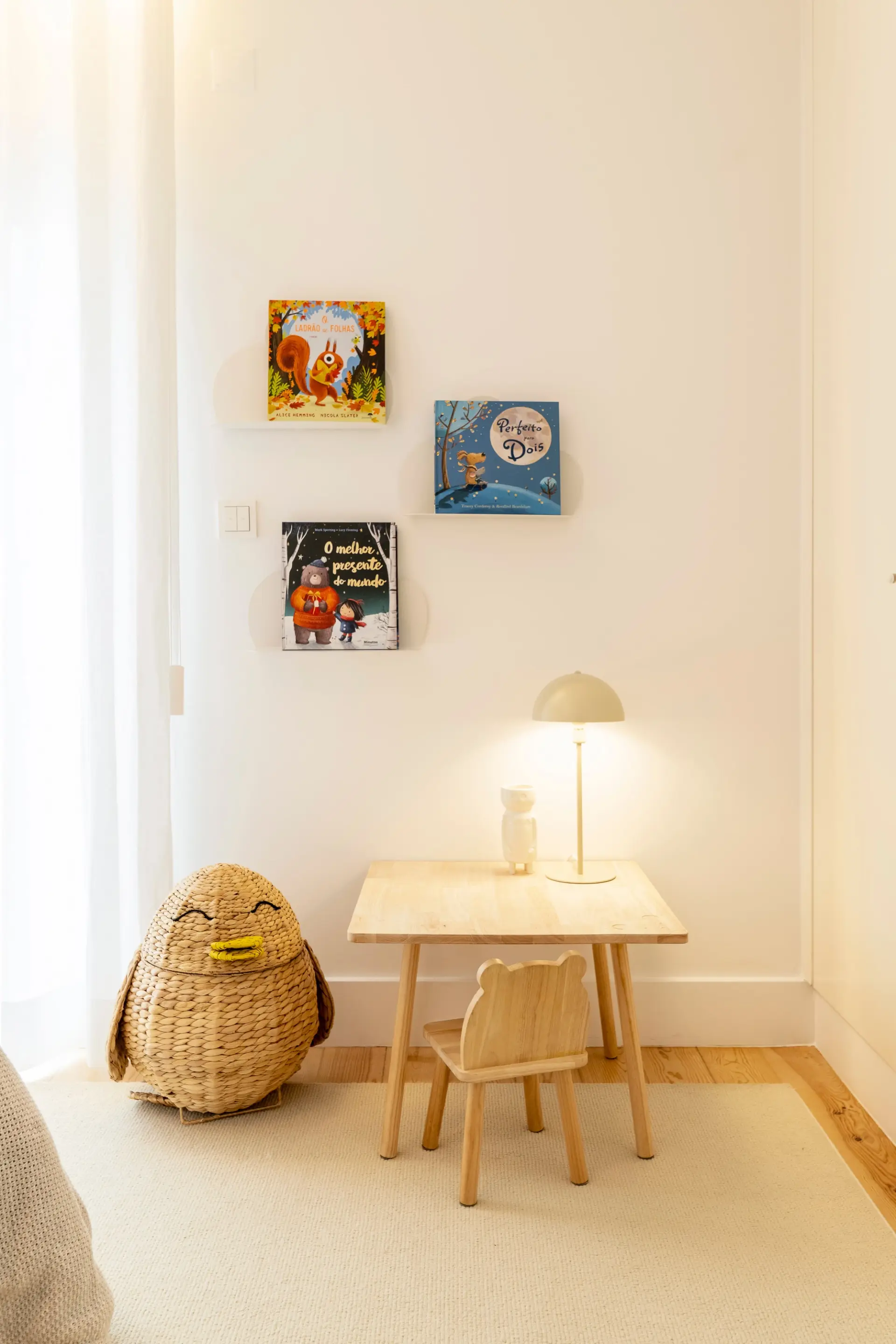 Zona de leitura infantil com mesa e cadeira em madeira, candeeiro e livros expostos na parede.