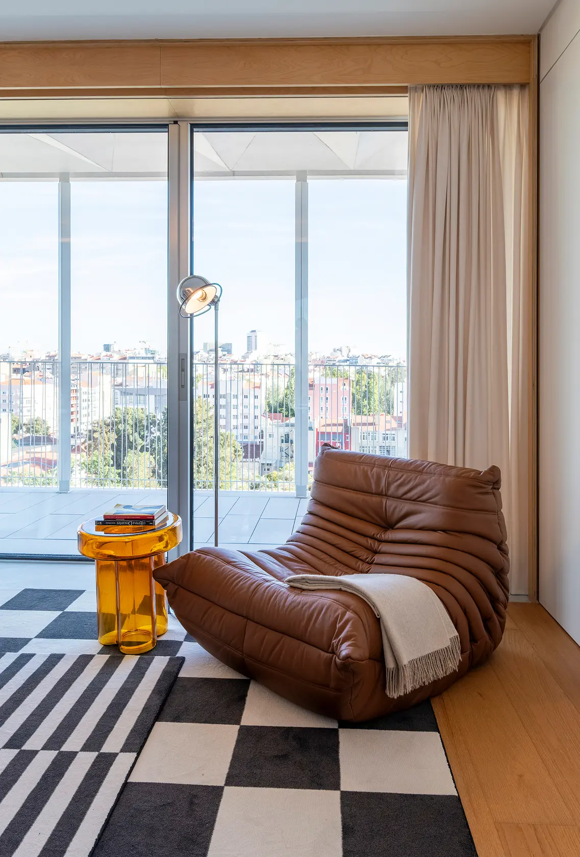 Cadeirão em pele de gomos cor cognac e mesa de apoio em vidro âmbar junto a janela com vista de cidade — quarto de hóspedes de luxo