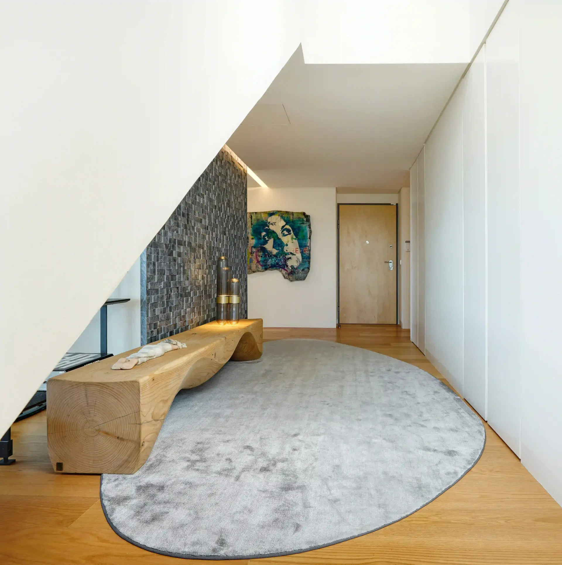 Hall de entrada minimalista com banco escultórico em madeira maciça, tapete oval cinza e parede revestida a pedra natural escura.