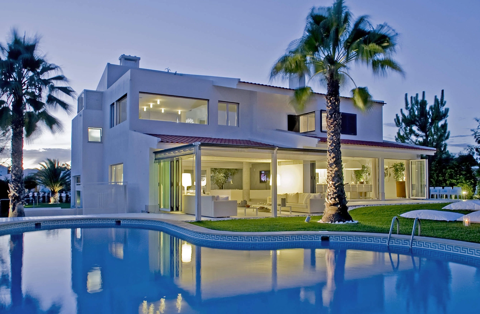 Moradia branca ao anoitecer com piscina, palmeiras e amplos envidraçados iluminados — design de interiores de luxo Tróia, Setúbal