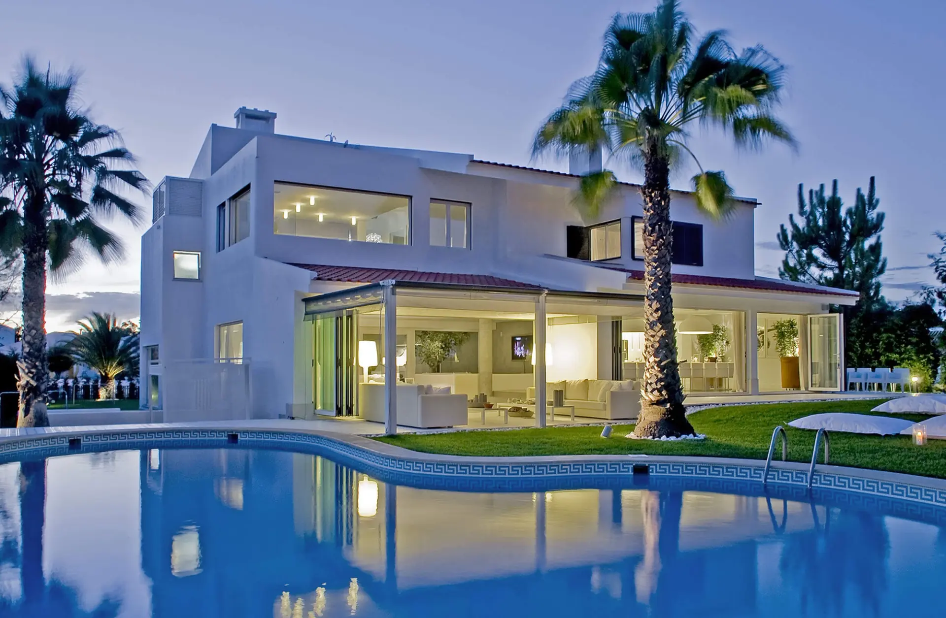 Moradia branca ao anoitecer com piscina, palmeiras e amplos envidraçados iluminados — design de interiores de luxo Tróia, Setúbal