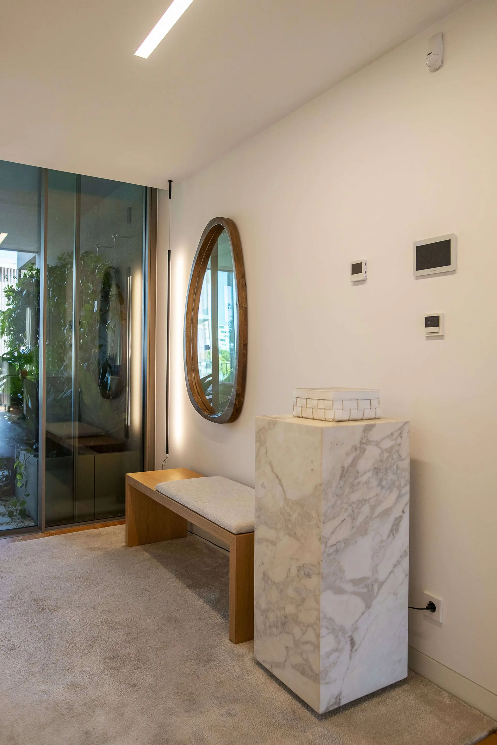 Round wood-framed mirror, oak bench and a Calacatta marble plinth with a woven box by a glass wall to the garden — luxury entrance hall