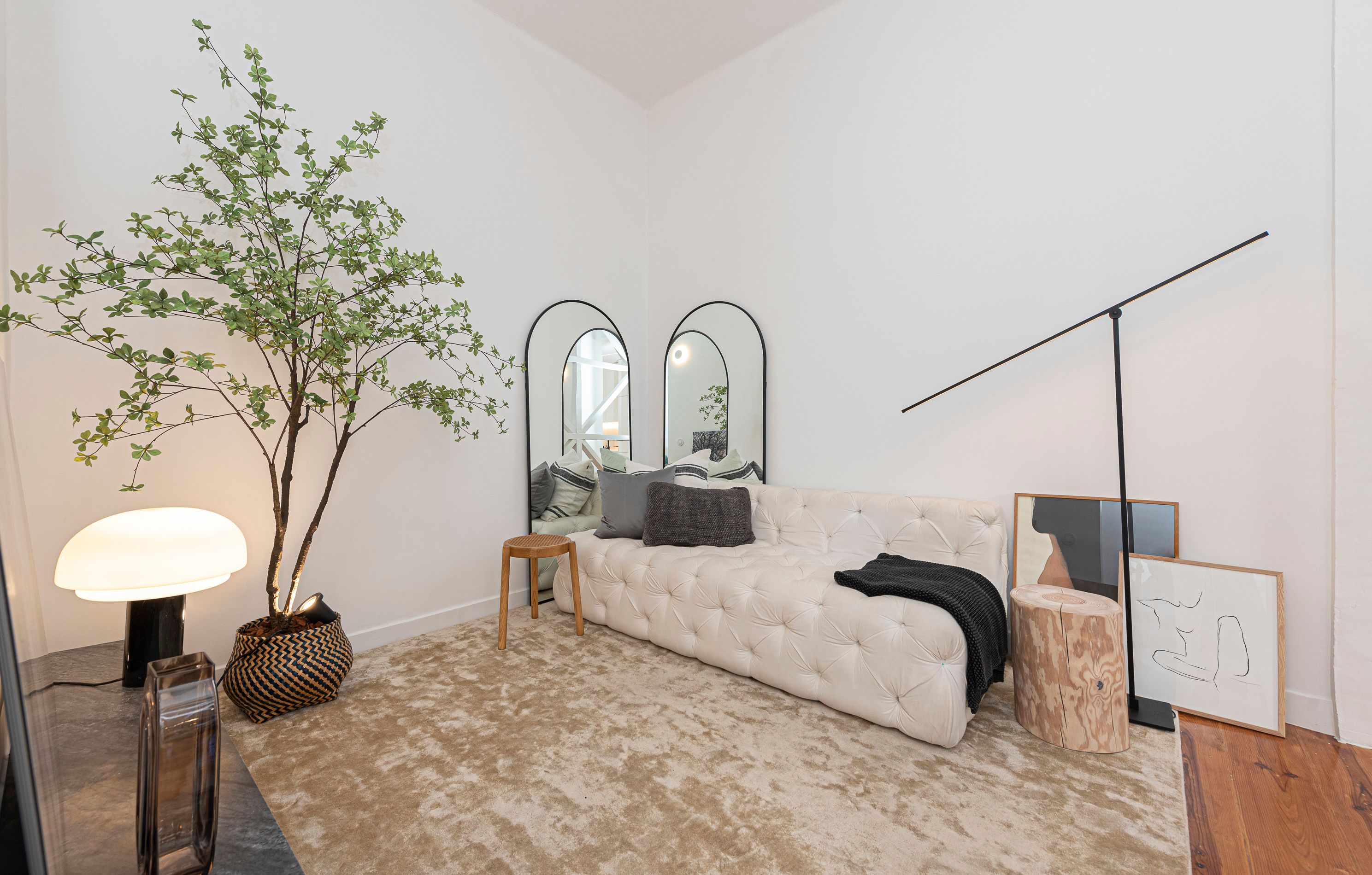 Living room with a white tufted sofa, arched mirrors, wooden side tables, a large potted tree and framed artwork leaning against the wall.