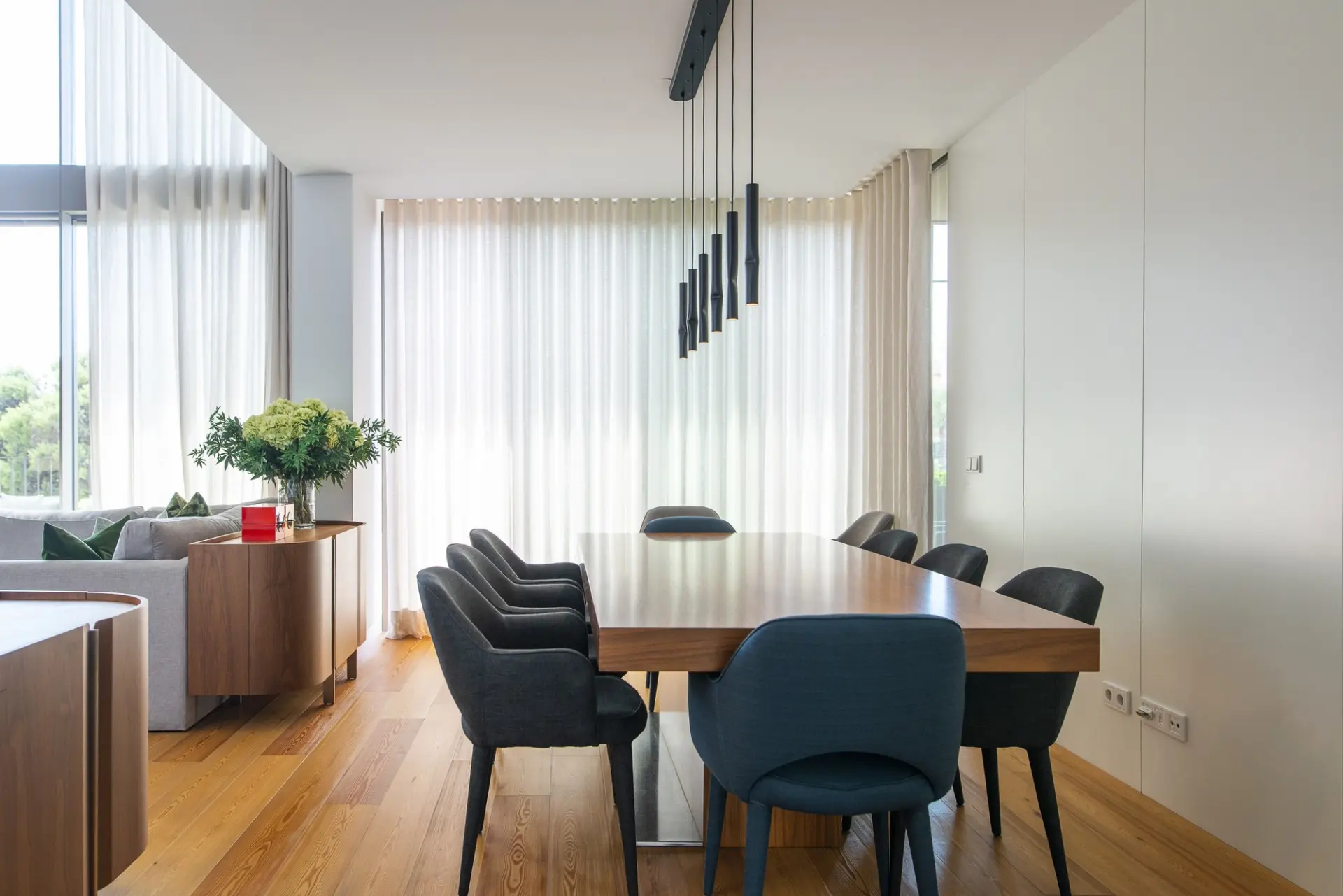 Dining room of the Cascais Apartment, with a wooden table, dark chairs, pendant lights and tall windows
