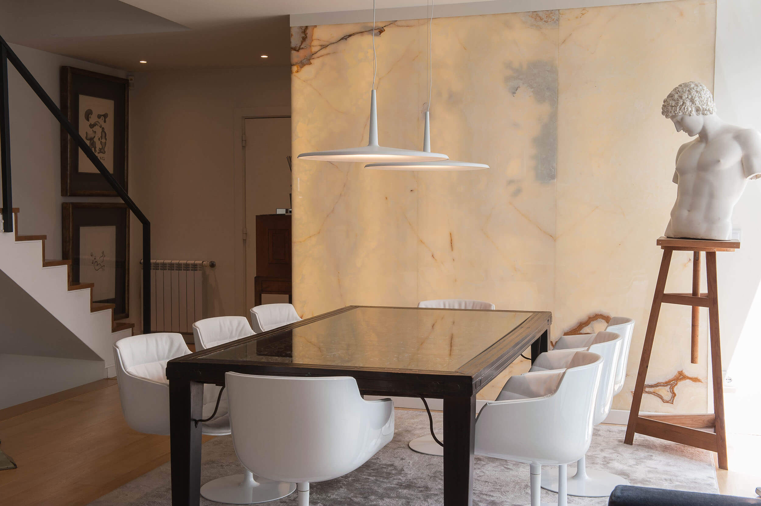 Dark-framed glass-top table, white leather swivel armchairs and a backlit onyx wall with a marble torso on a stand — luxury dining room