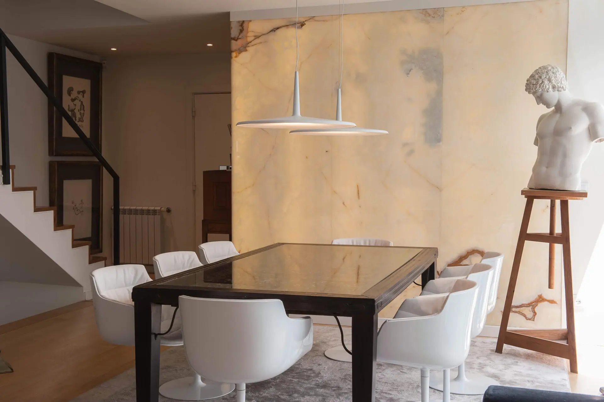 Dark-framed glass-top table, white leather swivel armchairs and a backlit onyx wall with a marble torso on a stand — luxury dining room