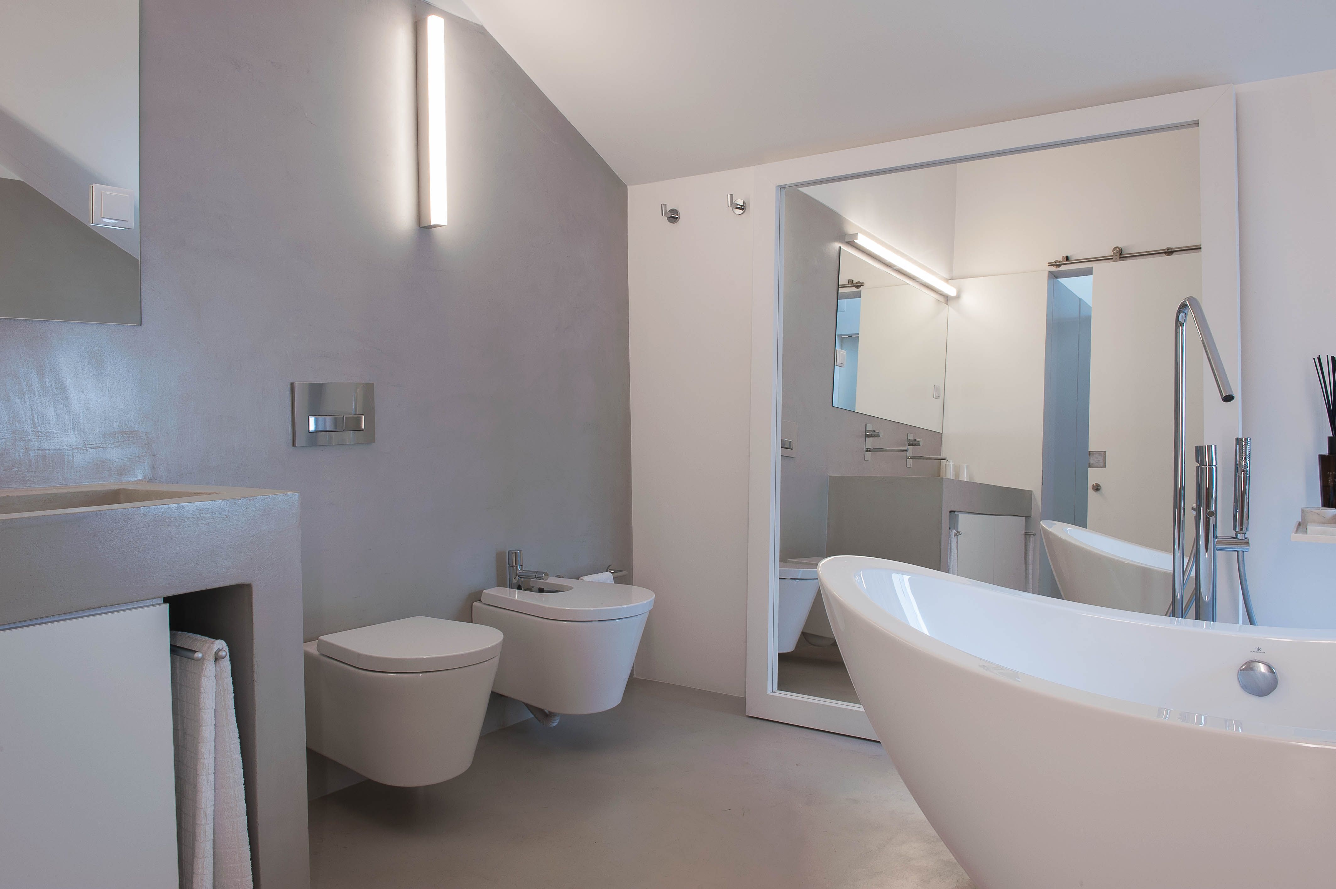Casa de banho em tons de cinzento e branco, com banheira independente, sanitários suspensos e espelho de grandes dimensões.