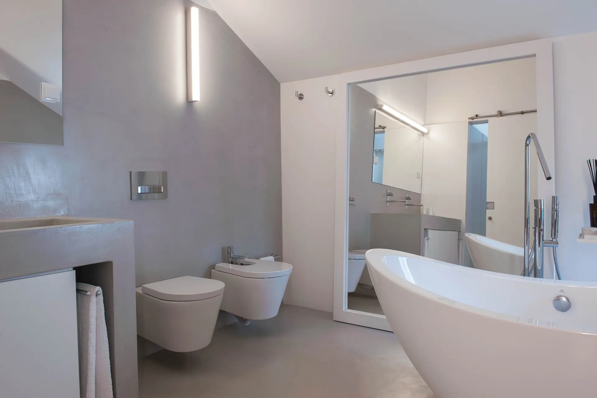 Casa de banho em tons de cinzento e branco, com banheira independente, sanitários suspensos e espelho de grandes dimensões.