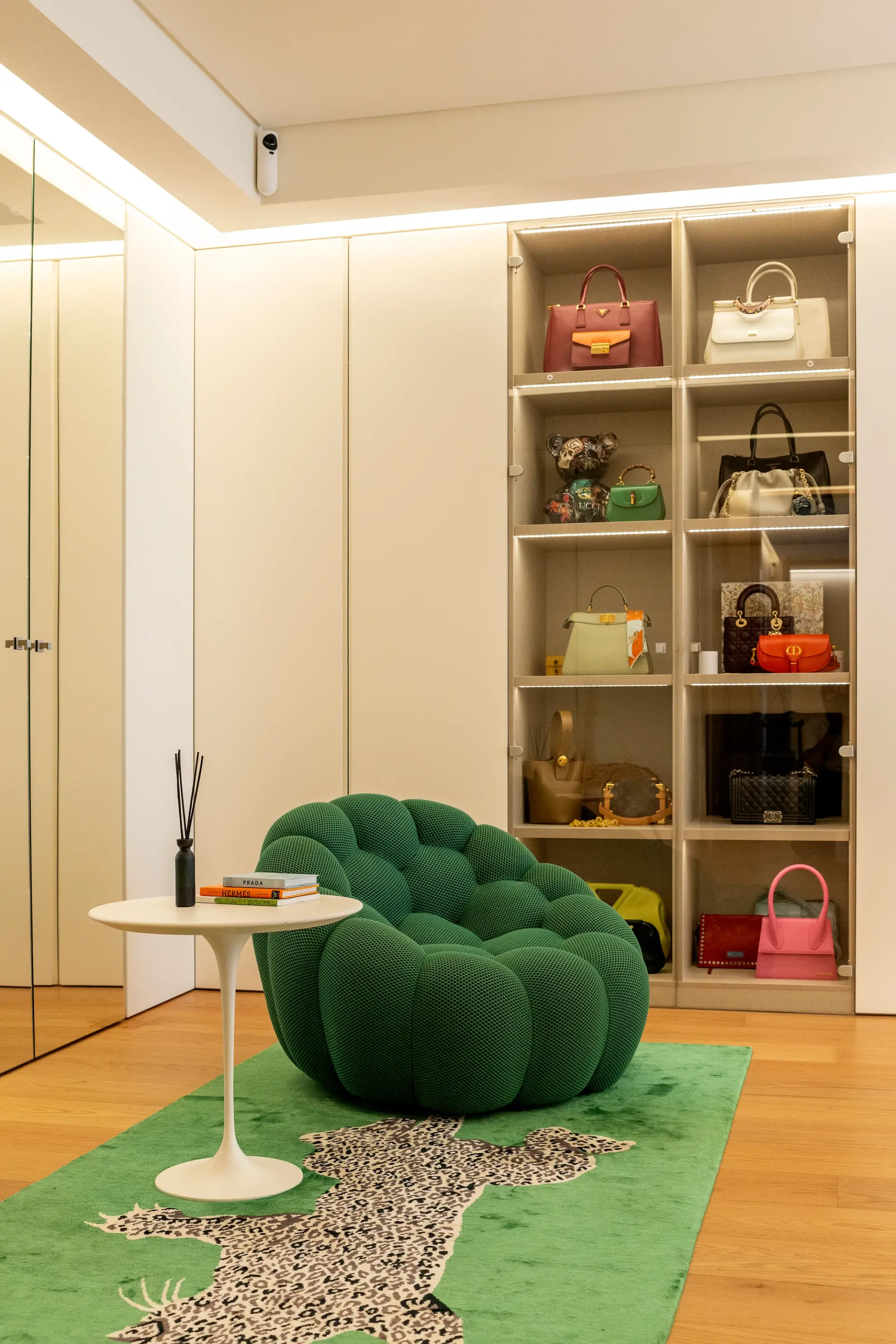 Closet com poltrona verde, mesa de apoio branca e armários com portas de vidro e iluminação interior.