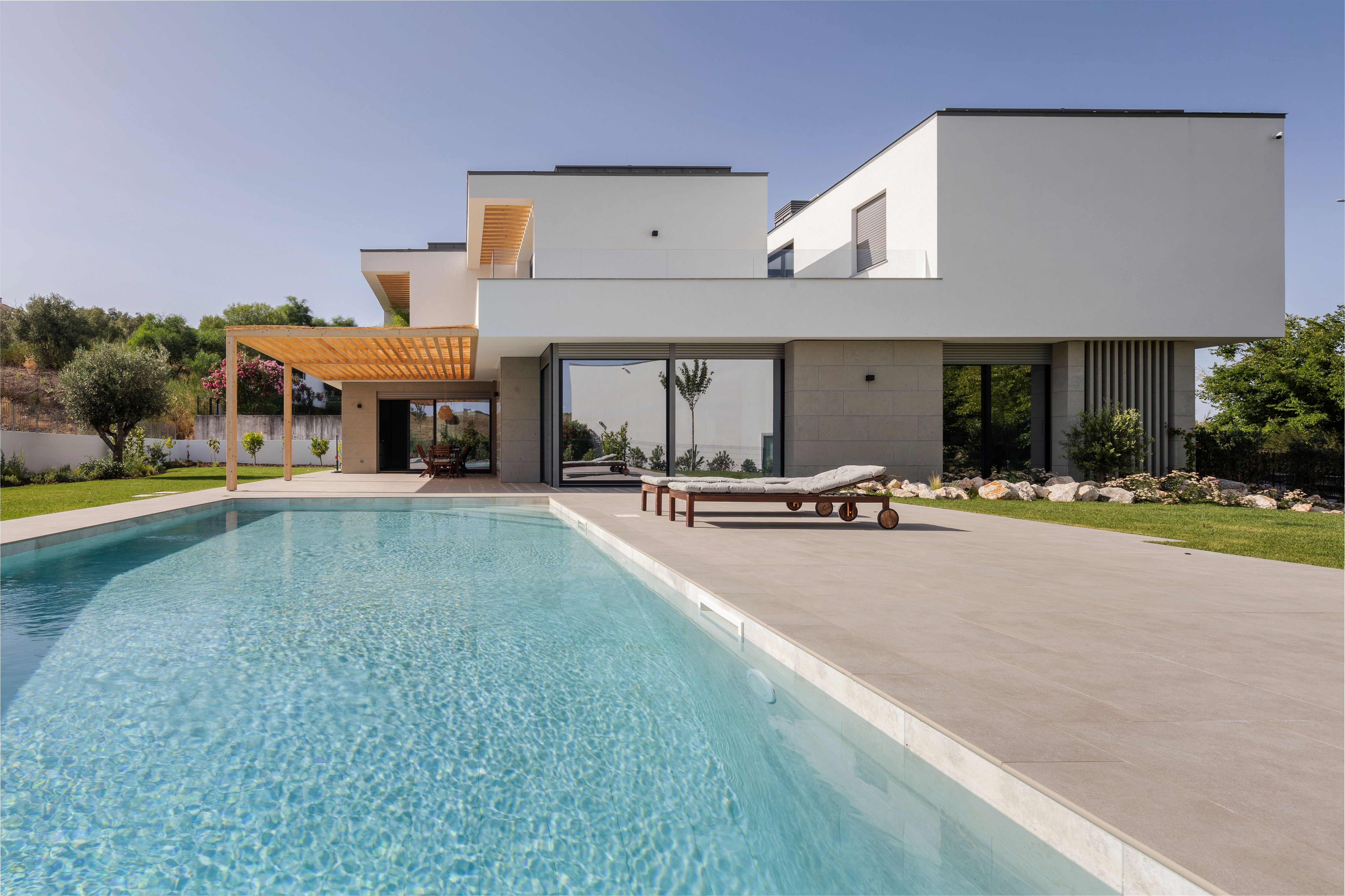 Moradia contemporânea com grandes janelas, piscina, espreguiçadeiras e zona de refeições sob pérgola de madeira.