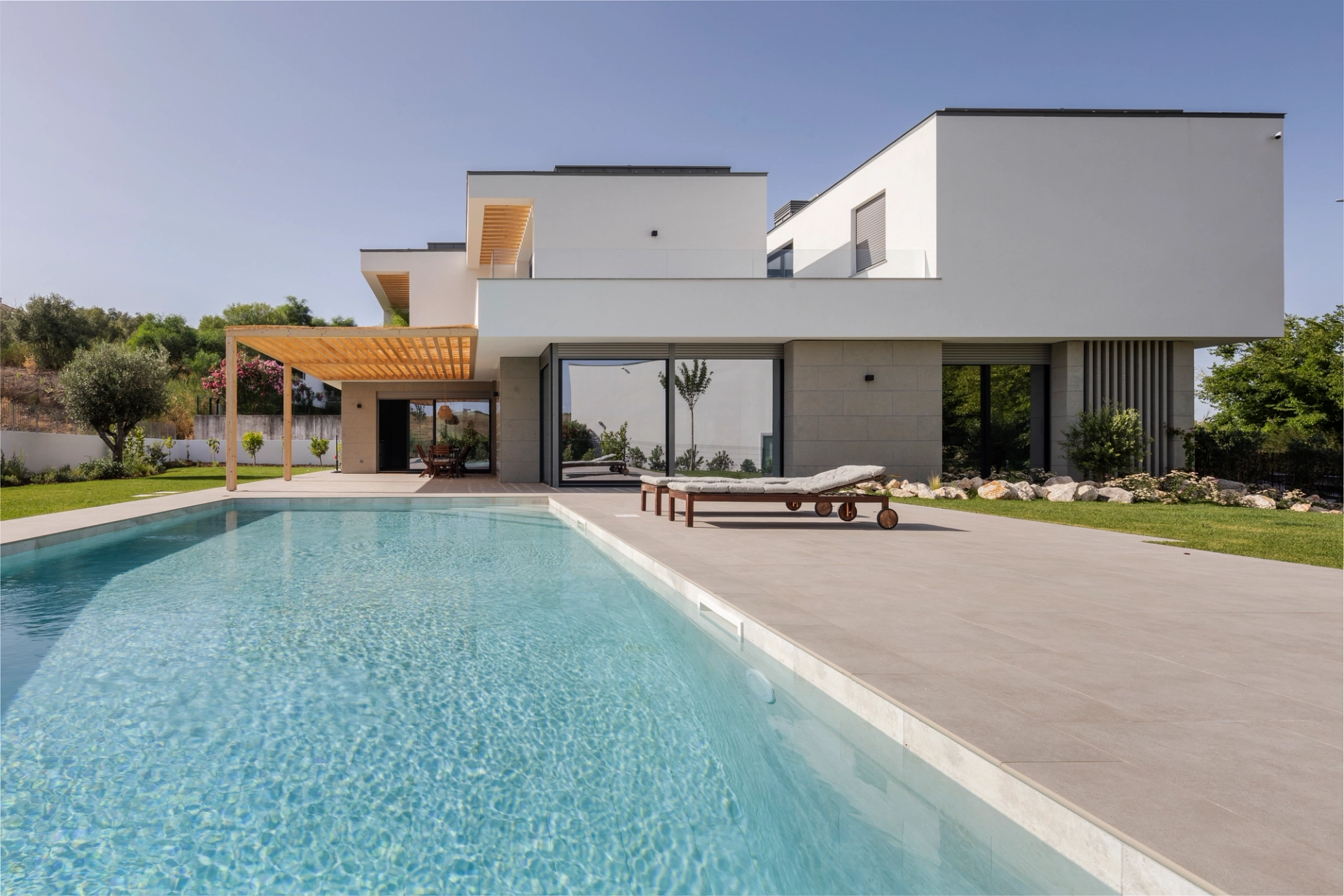 Moradia contemporânea com grandes janelas, piscina, espreguiçadeiras e zona de refeições sob pérgola de madeira.