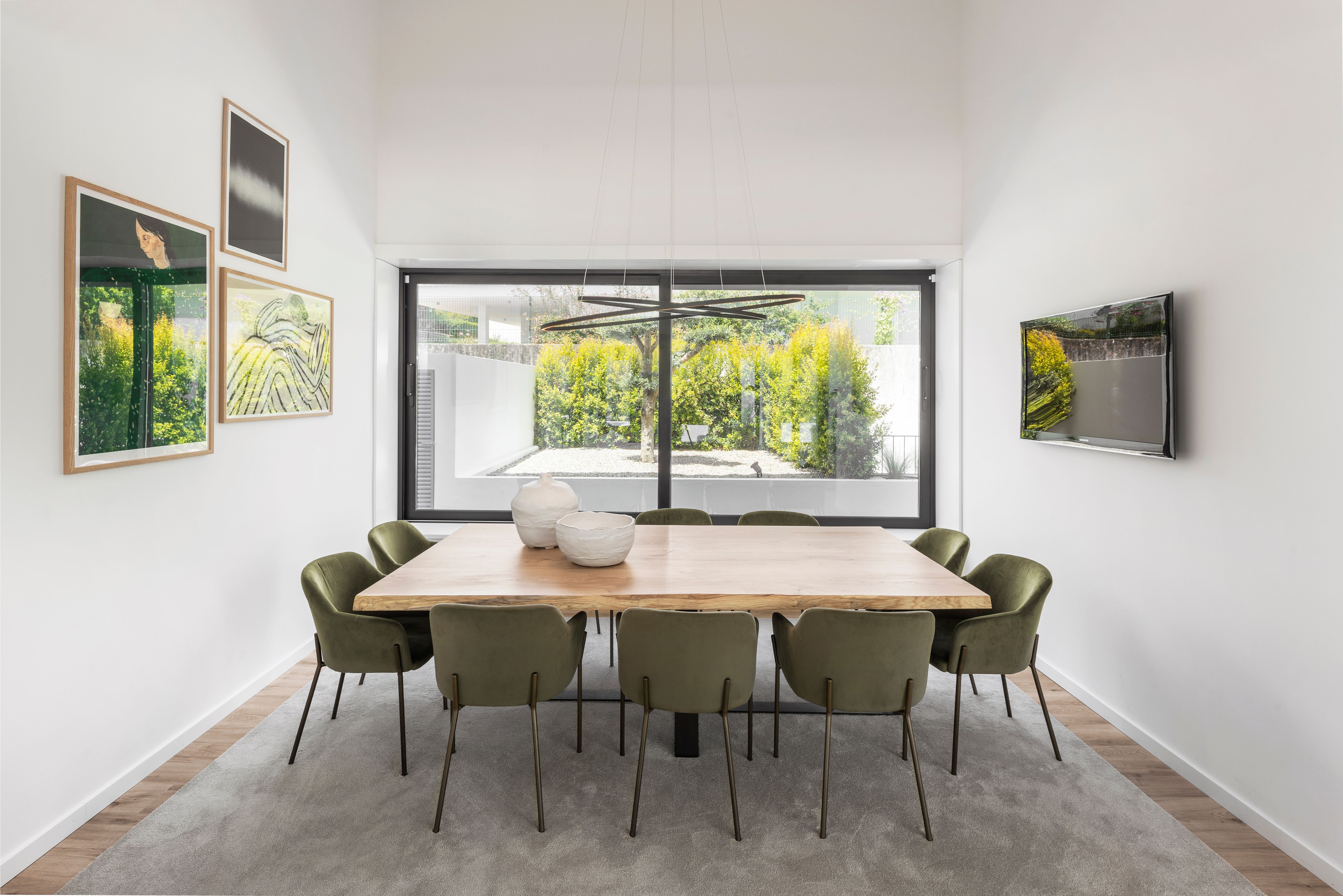 Sala de jantar com mesa retangular em madeira, cadeiras verdes, candeeiro suspenso e janela ampla com vista para o jardim.