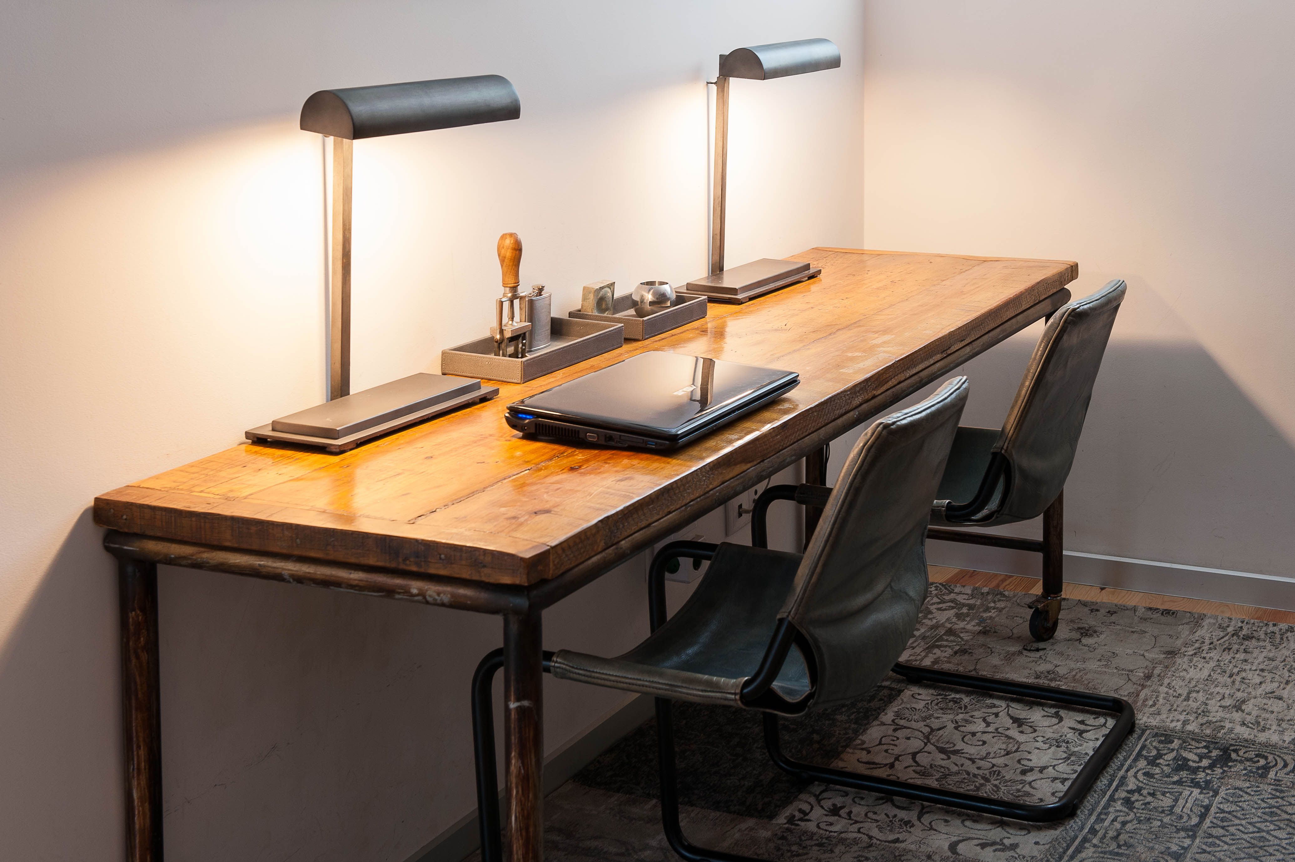 Zona de trabalho com secretária de madeira, dois candeeiros de mesa metálicos e duas cadeiras escuras sobre tapete com padrão.