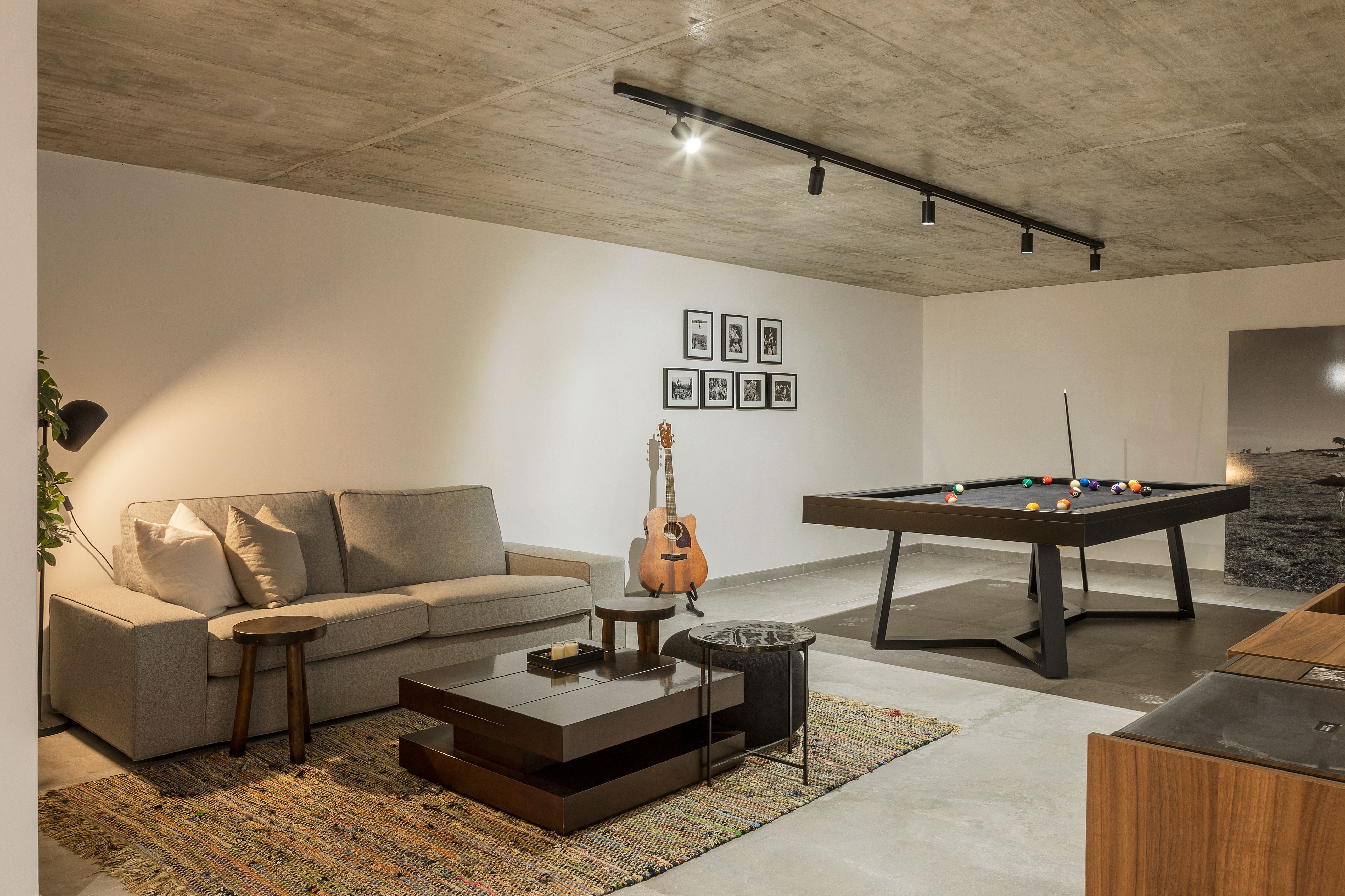 Sala de jogos com mesa de bilhar, sofá cinzento, mesas de centro em madeira e teto em betão à vista com iluminação em calha.