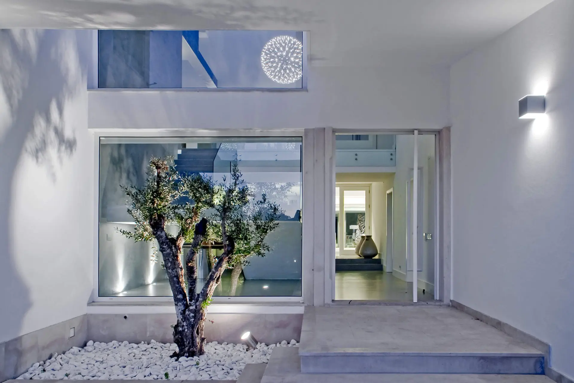 Entrada de moradia branca com oliveira em seixos e porta pivotante envidraçada — design de interiores de luxo Tróia, Setúbal