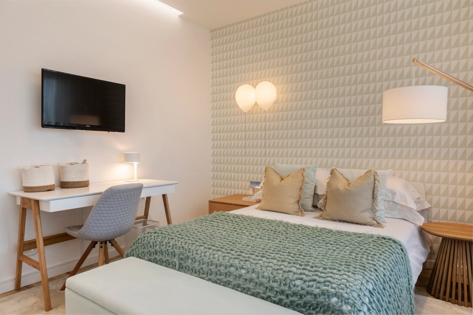Quarto com cama de casal, manta verde, papel de parede geométrico, secretária e iluminação decorativa.