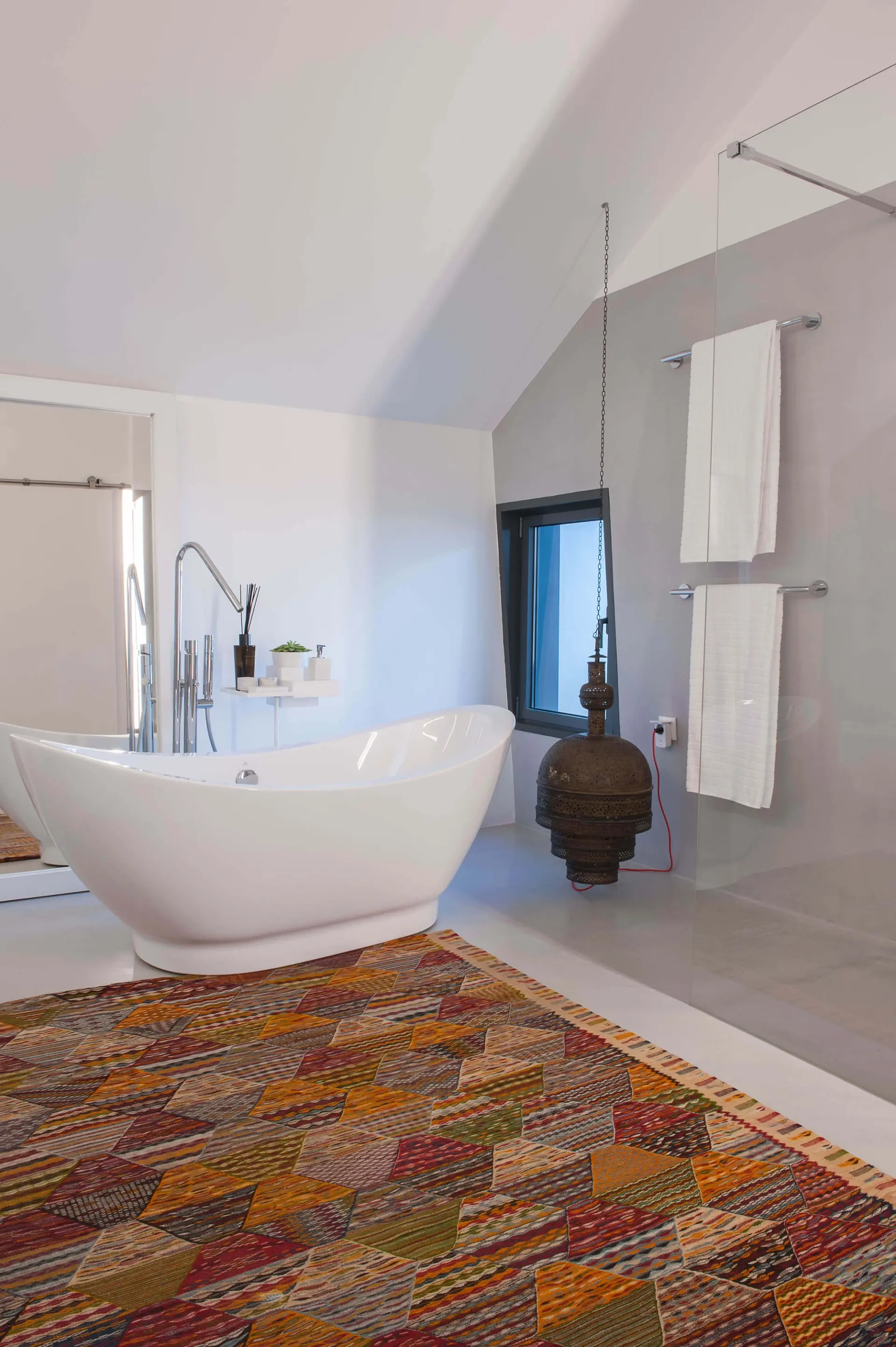 White oval freestanding tub, a multicolour kilim rug, a hammered-brass pendant and a grey microcement wall — luxury bathroom