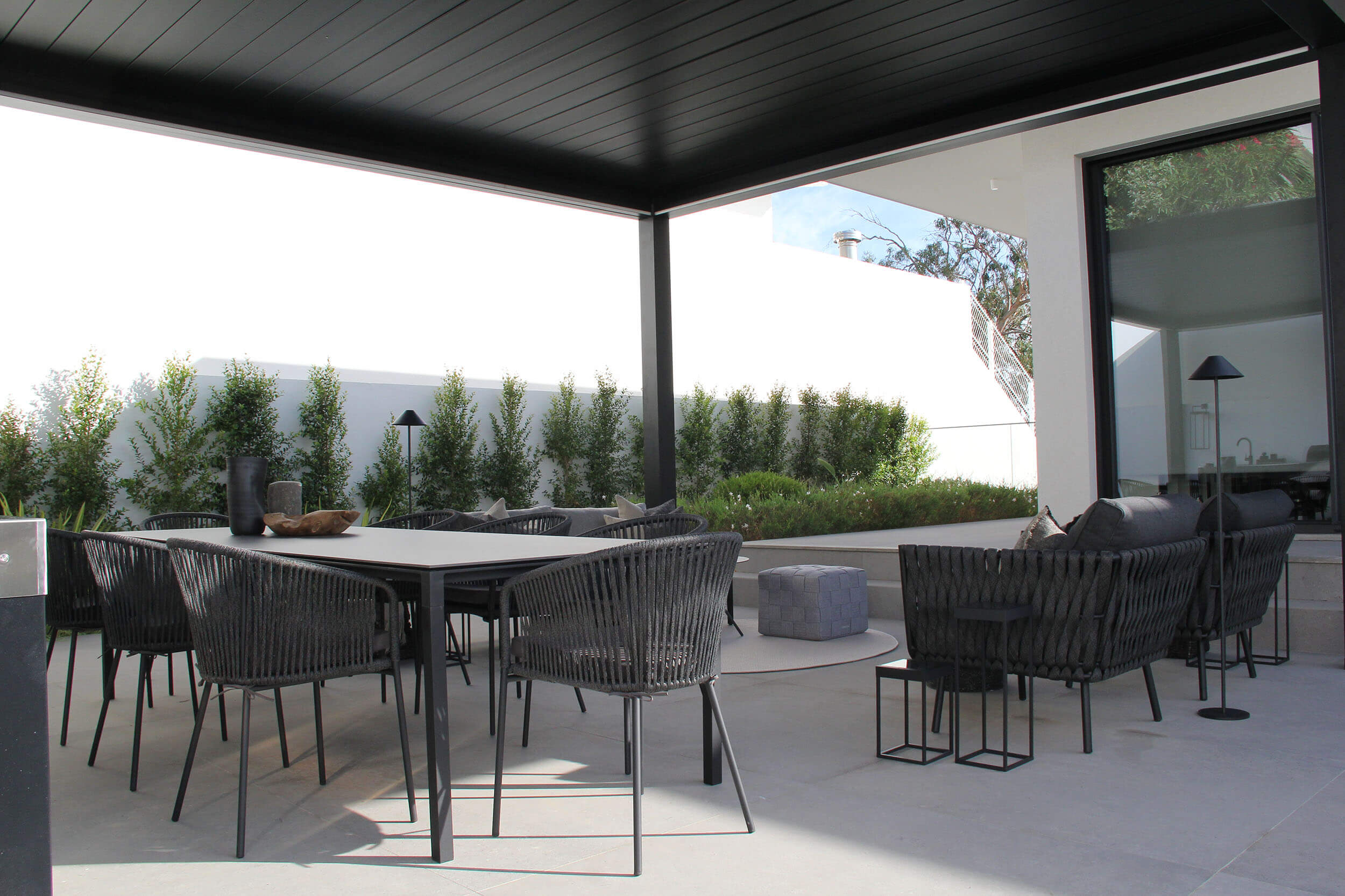 Mesa de refeição e cadeiras em corda escura sob pérgola preta, sofás de exterior e sebe num terraço pavimentado — exterior de luxo