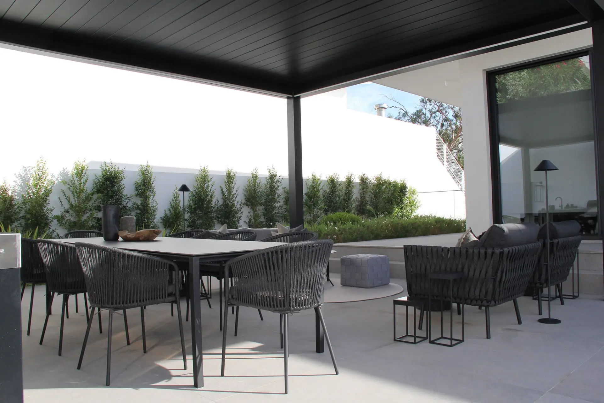 Mesa de refeição e cadeiras em corda escura sob pérgola preta, sofás de exterior e sebe num terraço pavimentado — exterior de luxo
