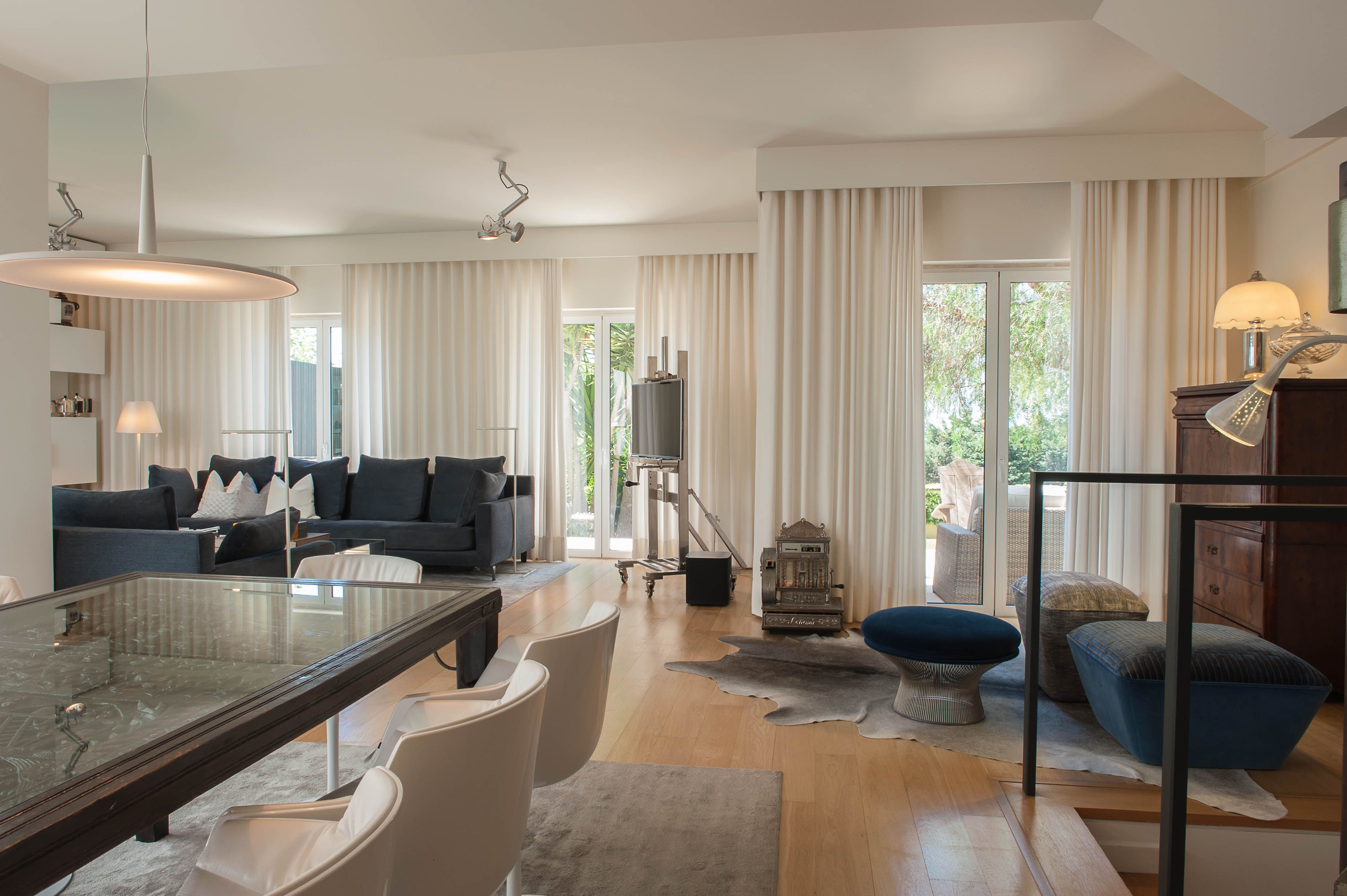 Contemporary open-plan living and dining room with dark sofas, white chairs and floor-to-ceiling curtains.