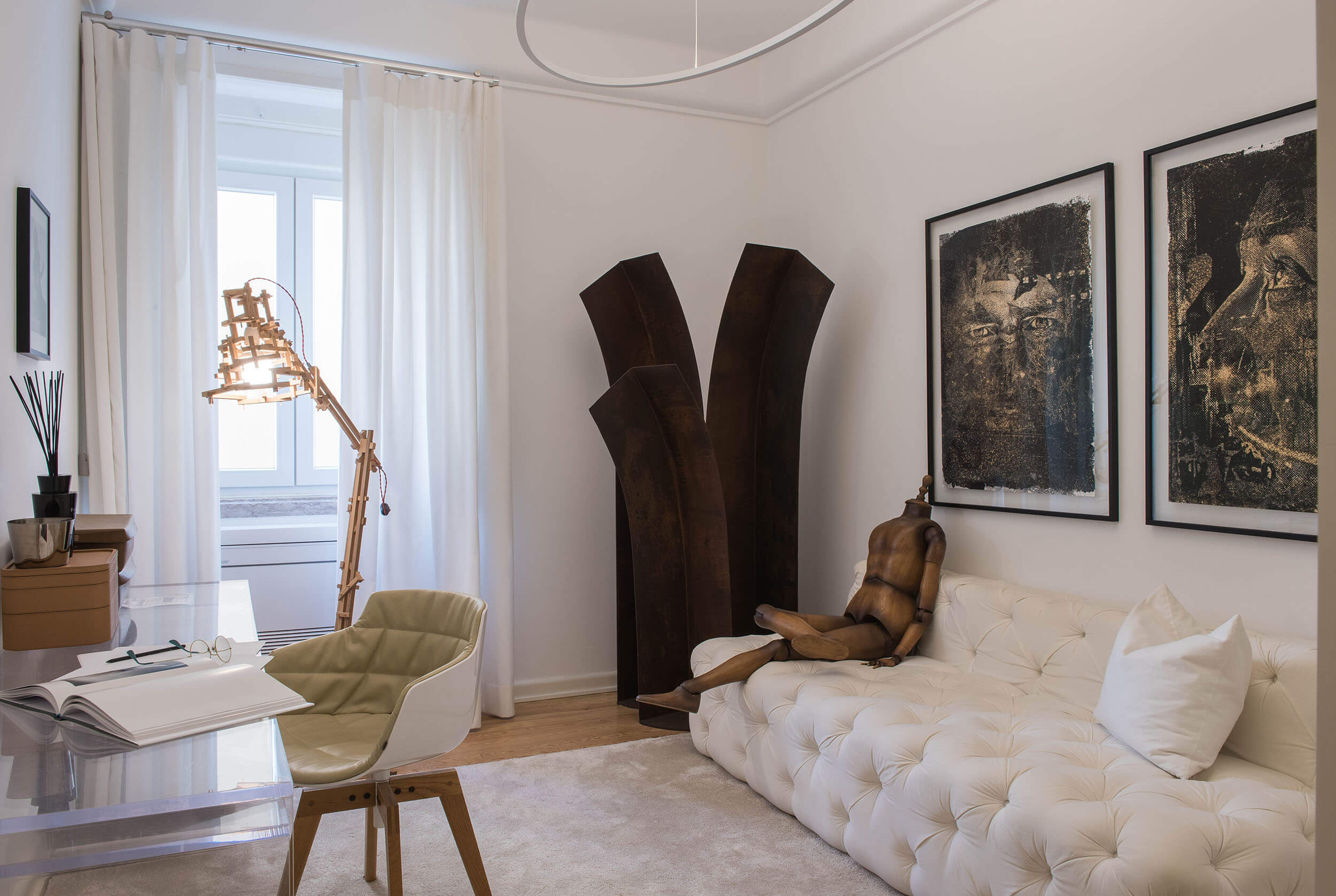 White tufted sofa, articulated wood floor lamp and corten steel sculpture beside framed portrait prints — luxury home office design
