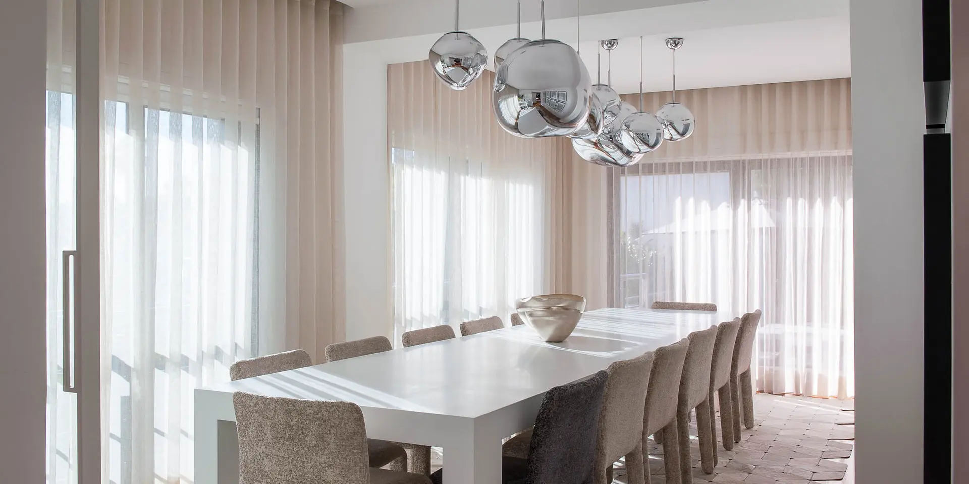 Sala de jantar moderna com mesa longa, cadeiras estofadas e iluminação suspensa. Exemplo de layout e coerência ao decorar casa nova
