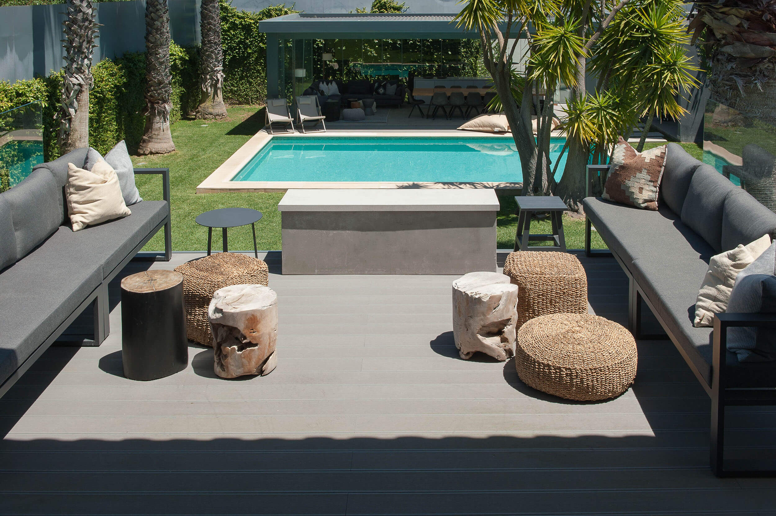 Dois sofás cinza modulares em deck composto, tocos de madeira e mesa em betão junto a piscina e palmeiras — exterior de luxo