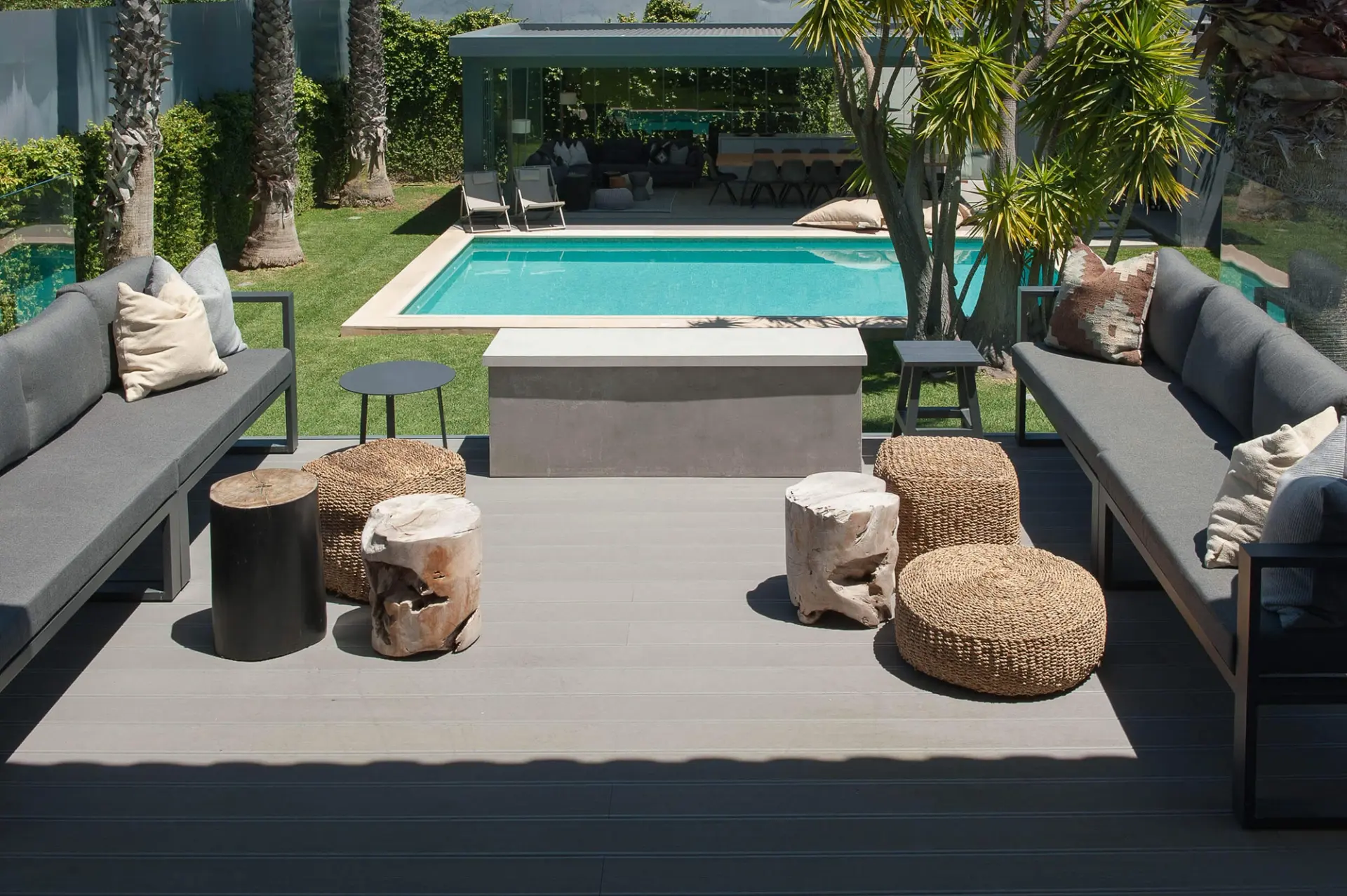 Dois sofás cinza modulares em deck composto, tocos de madeira e mesa em betão junto a piscina e palmeiras — exterior de luxo