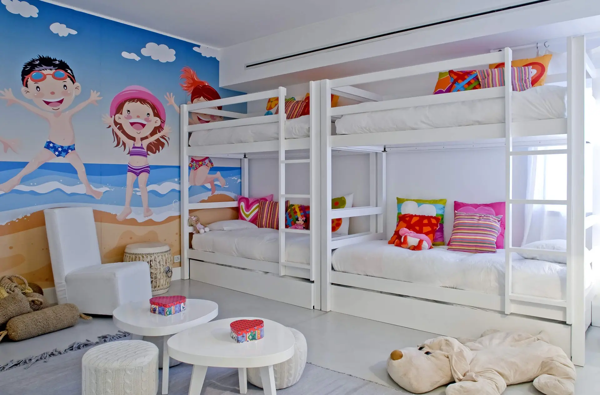 Quarto infantil com beliches brancos, almofadas coloridas e mural de praia ilustrado — design de interiores de luxo Tróia, Setúbal