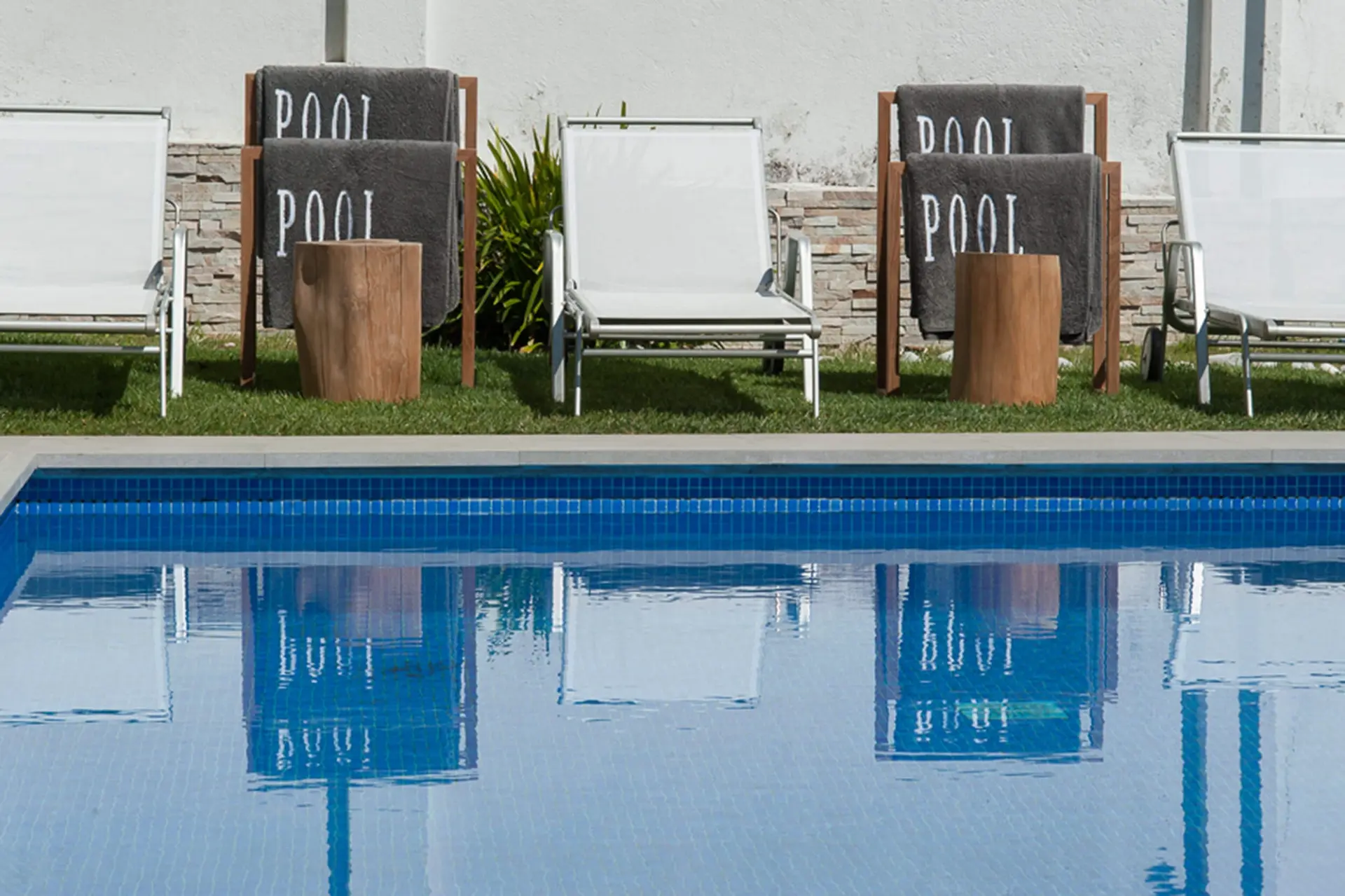 Espreguiçadeiras com toalhas bordadas 'POOL', tocos de madeira como mesa de apoio e piscina de mosaico azul — exterior de luxo