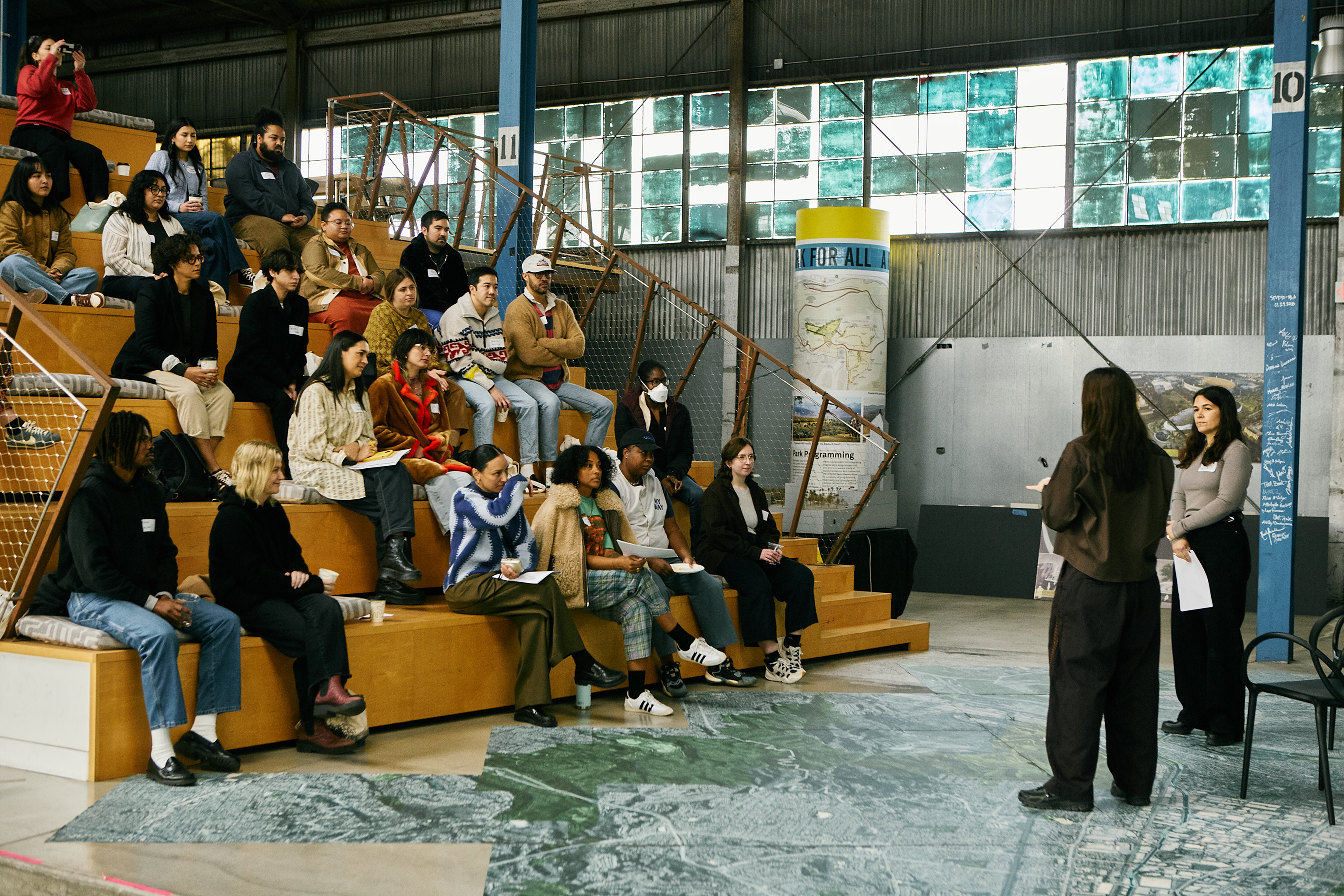 Fellows seated on bleachers listen to two facilitators in a converted warehouse.