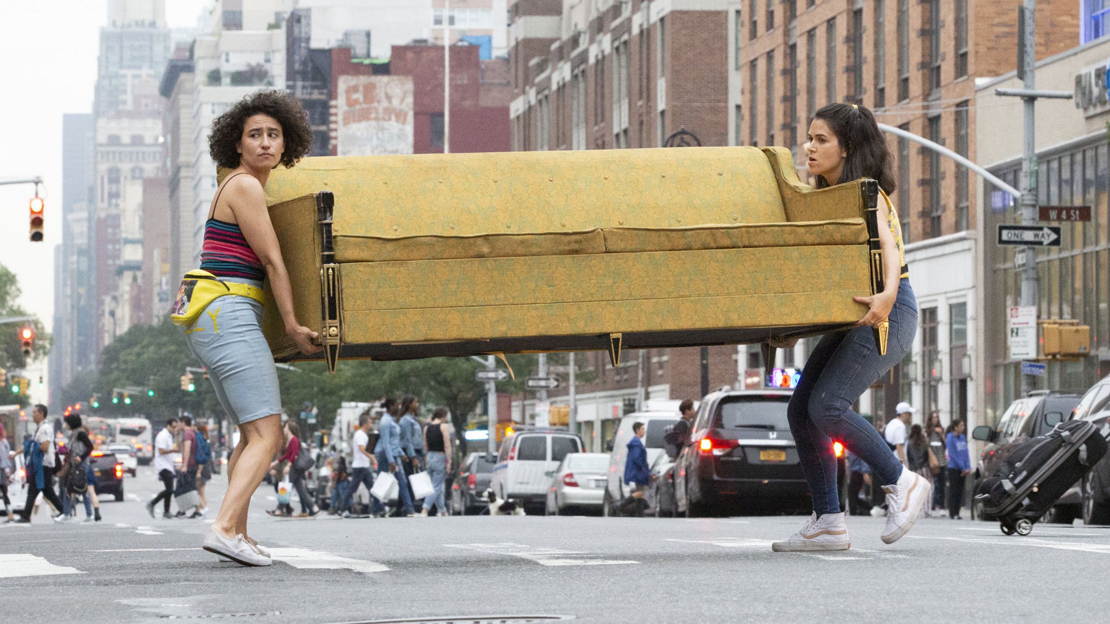 Abbi and Ilana carry a sofa across a bustling NY intersection.