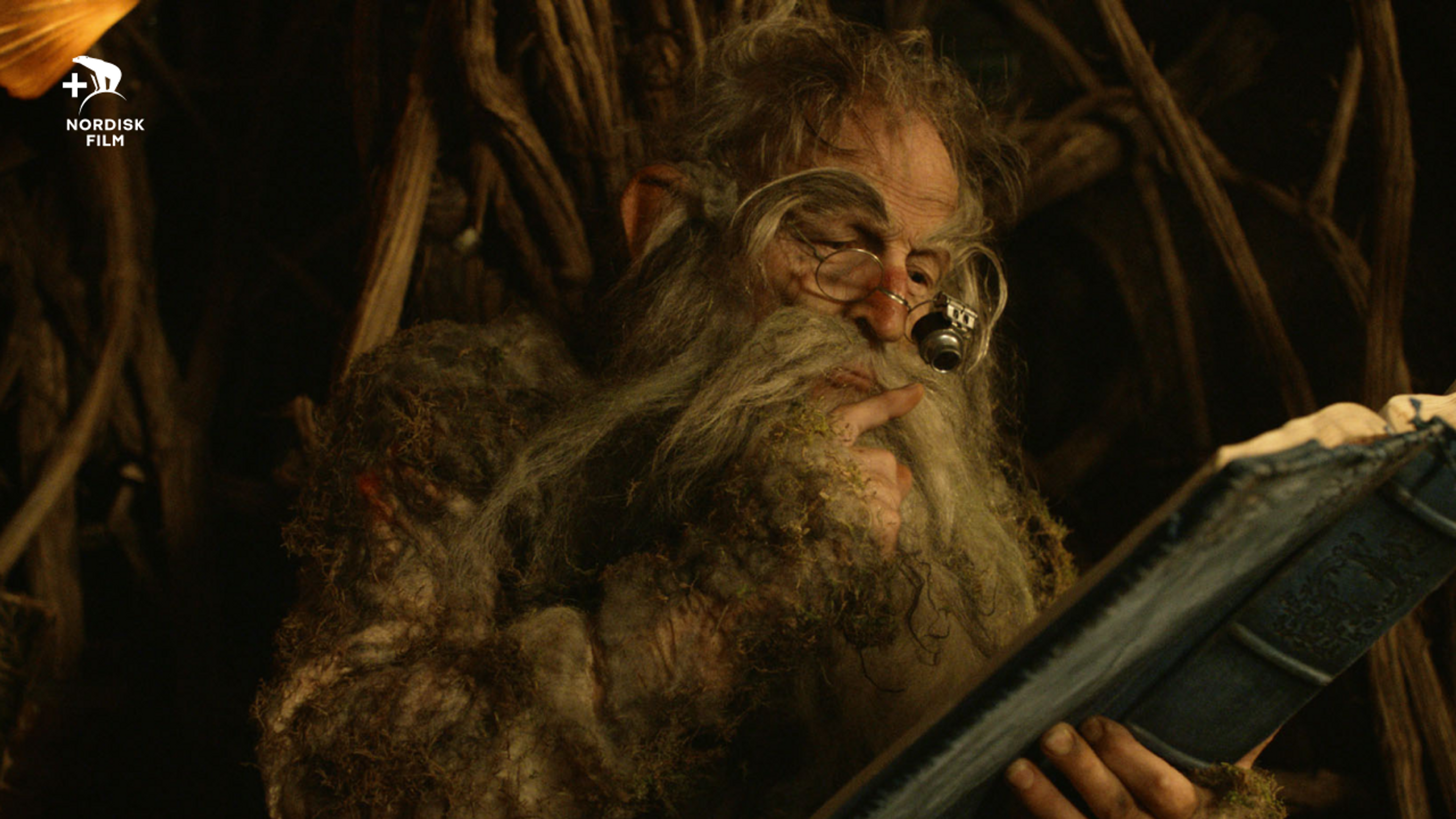 En gammel nisse sitter i en mørk grotte og studerer en ønskeliste fra barna