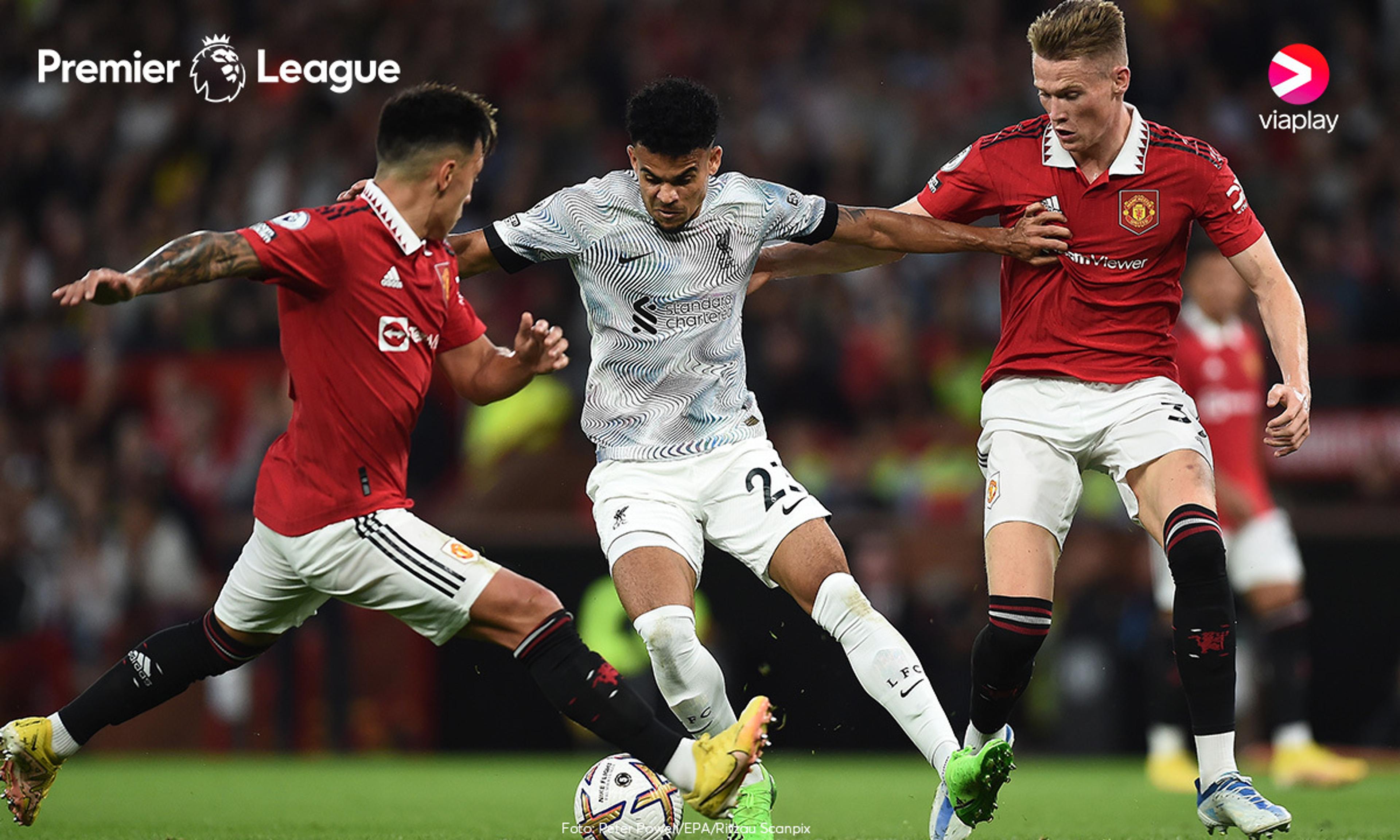 Tre spillere, to i rødt, en i hvitt, kjemper hard om ballen med en fullpakket stadion i bakgrunnen.