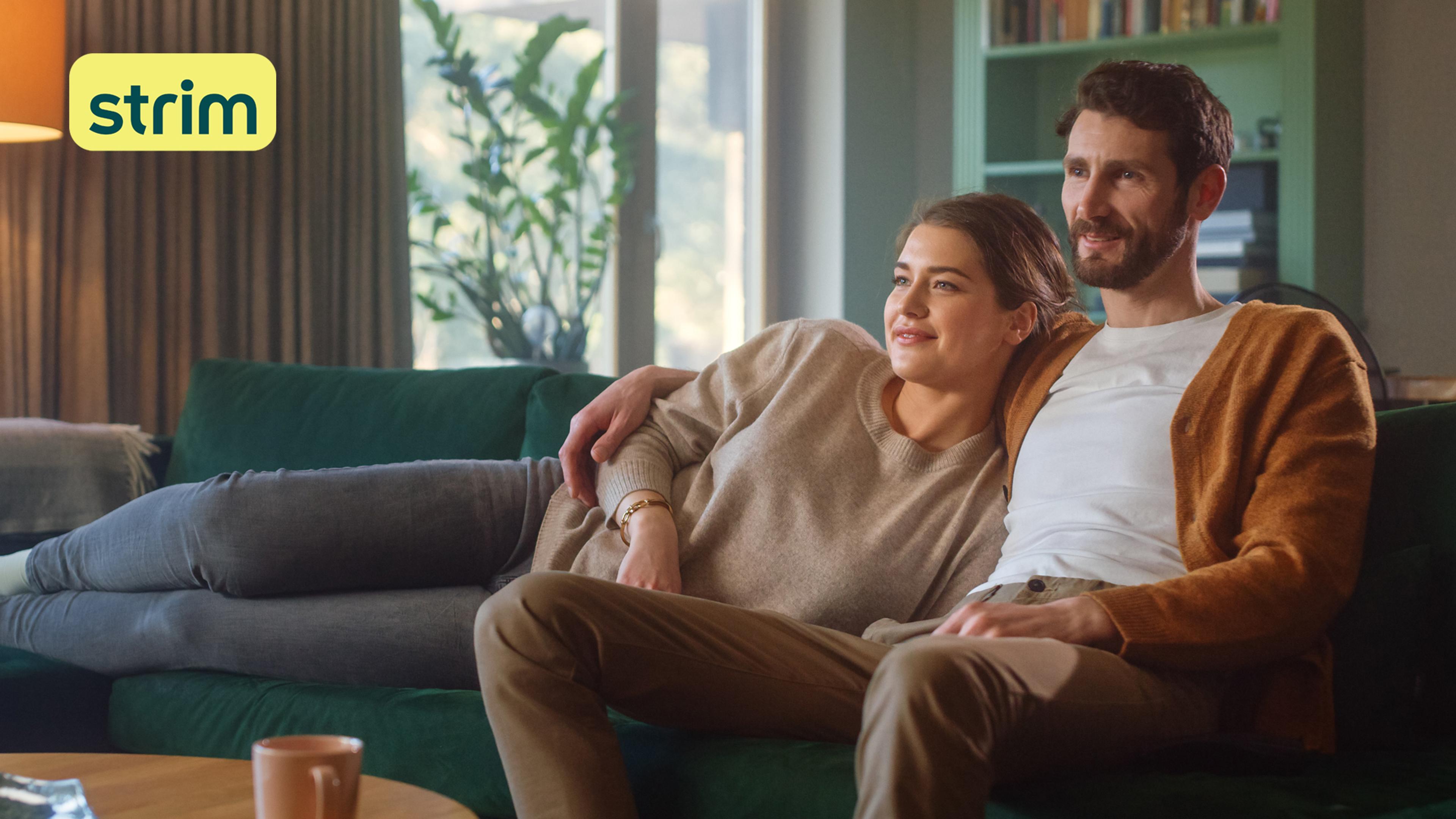 Par sitter i grønn sofa og ser på TV.