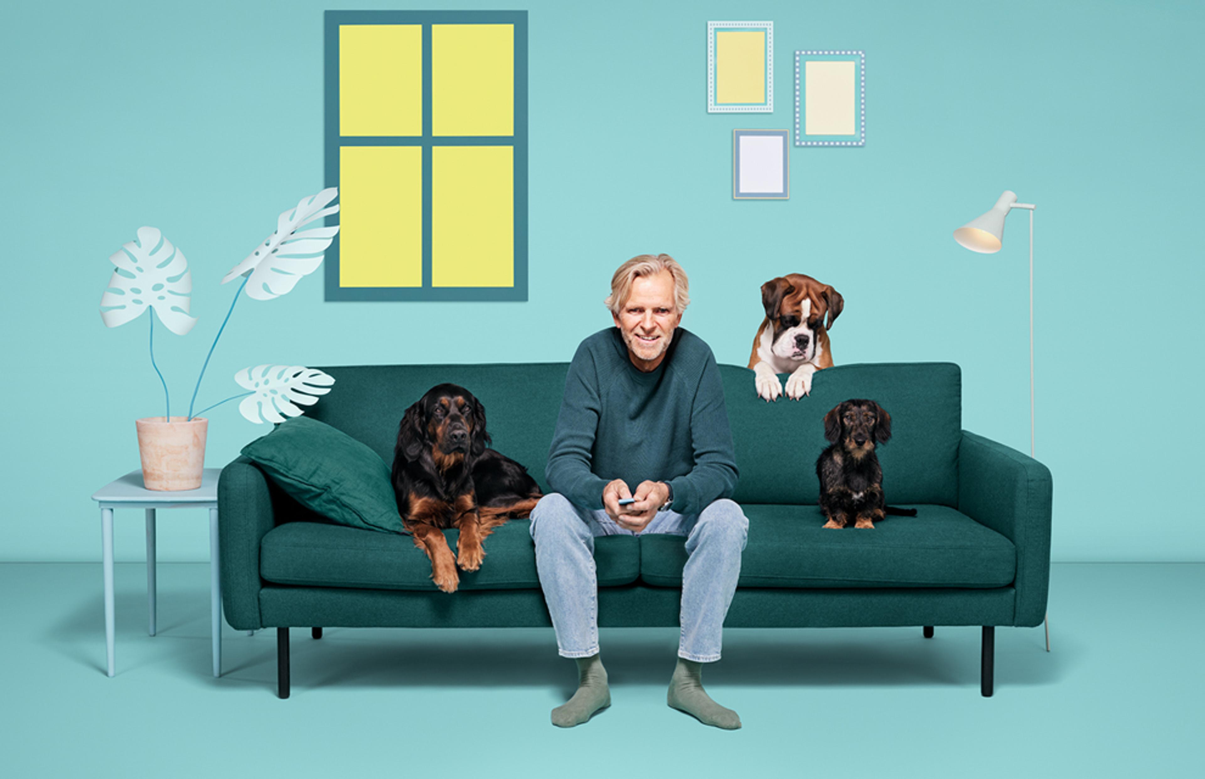 Eldre mann sitter fornøyd i sofaen og ser på TV med tre hunder som følger spent med på det som skjer.
