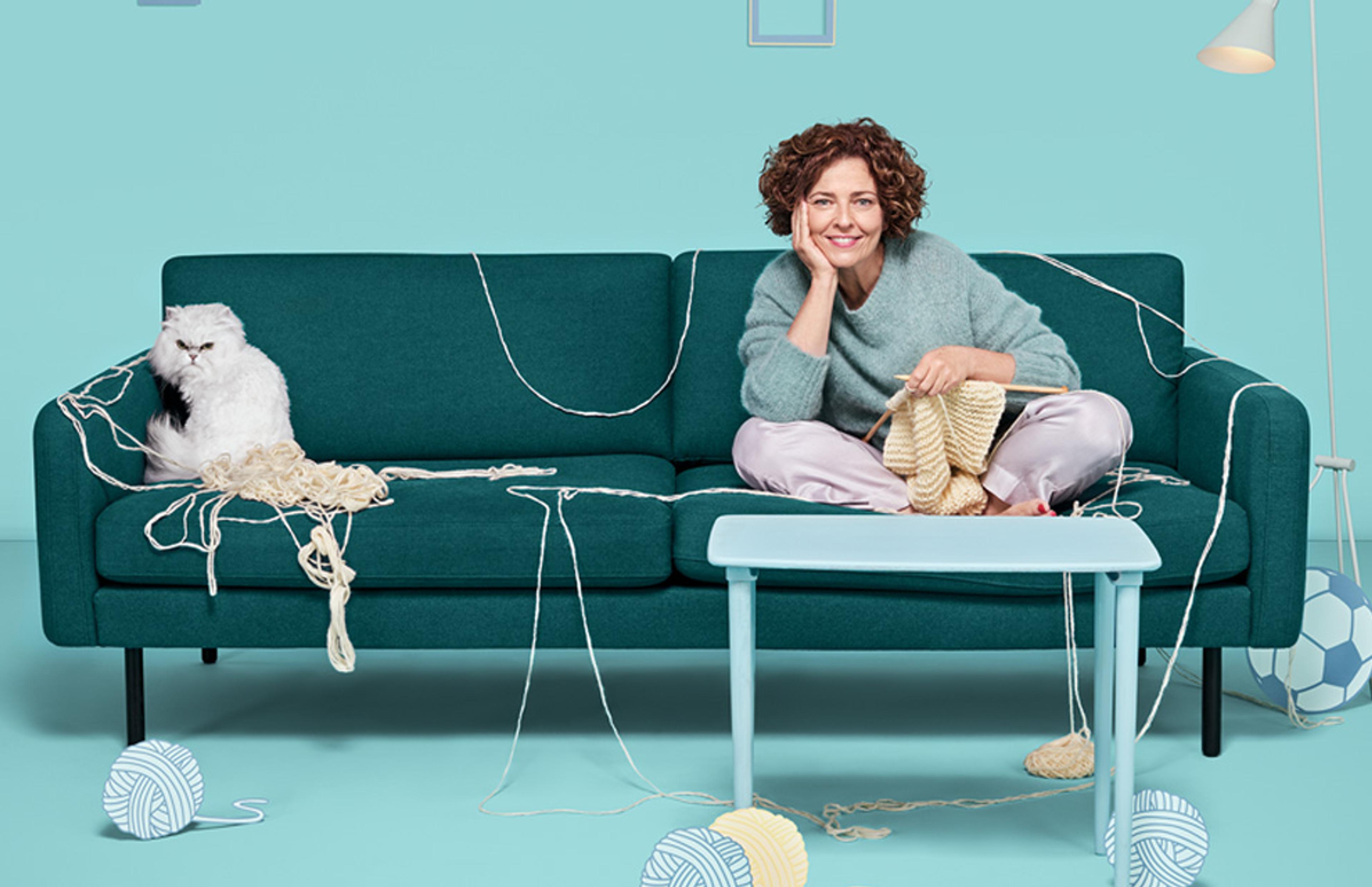 Kvinne ser TV med Strim mens hun strikker. Misfornøyd katt sitter på sofaen med kvinnen og har dratt garn over hele sofaen.