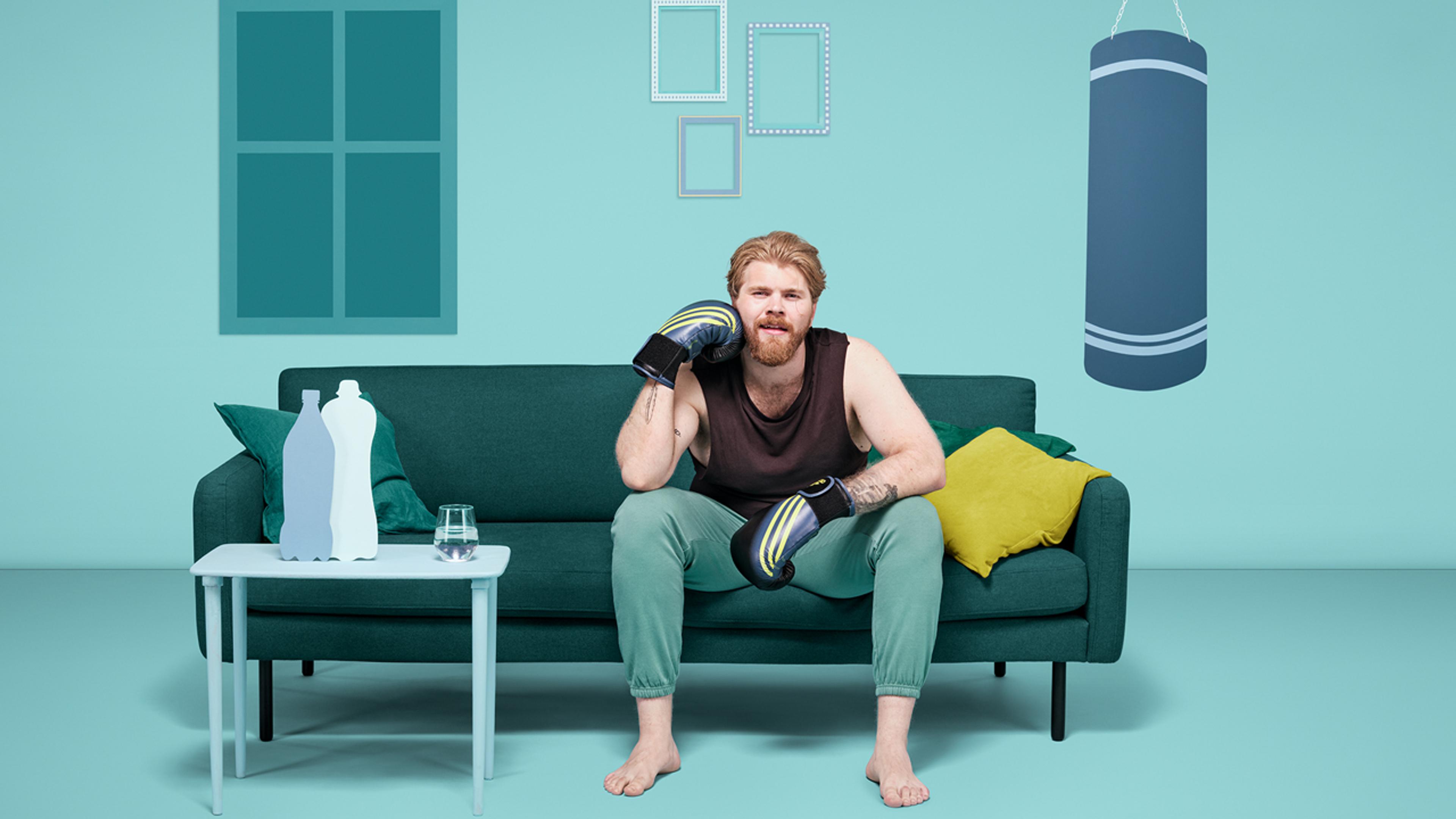 Mann med boksehansker og treningsklær sitter på sofa i turkis rom og ser på TV.